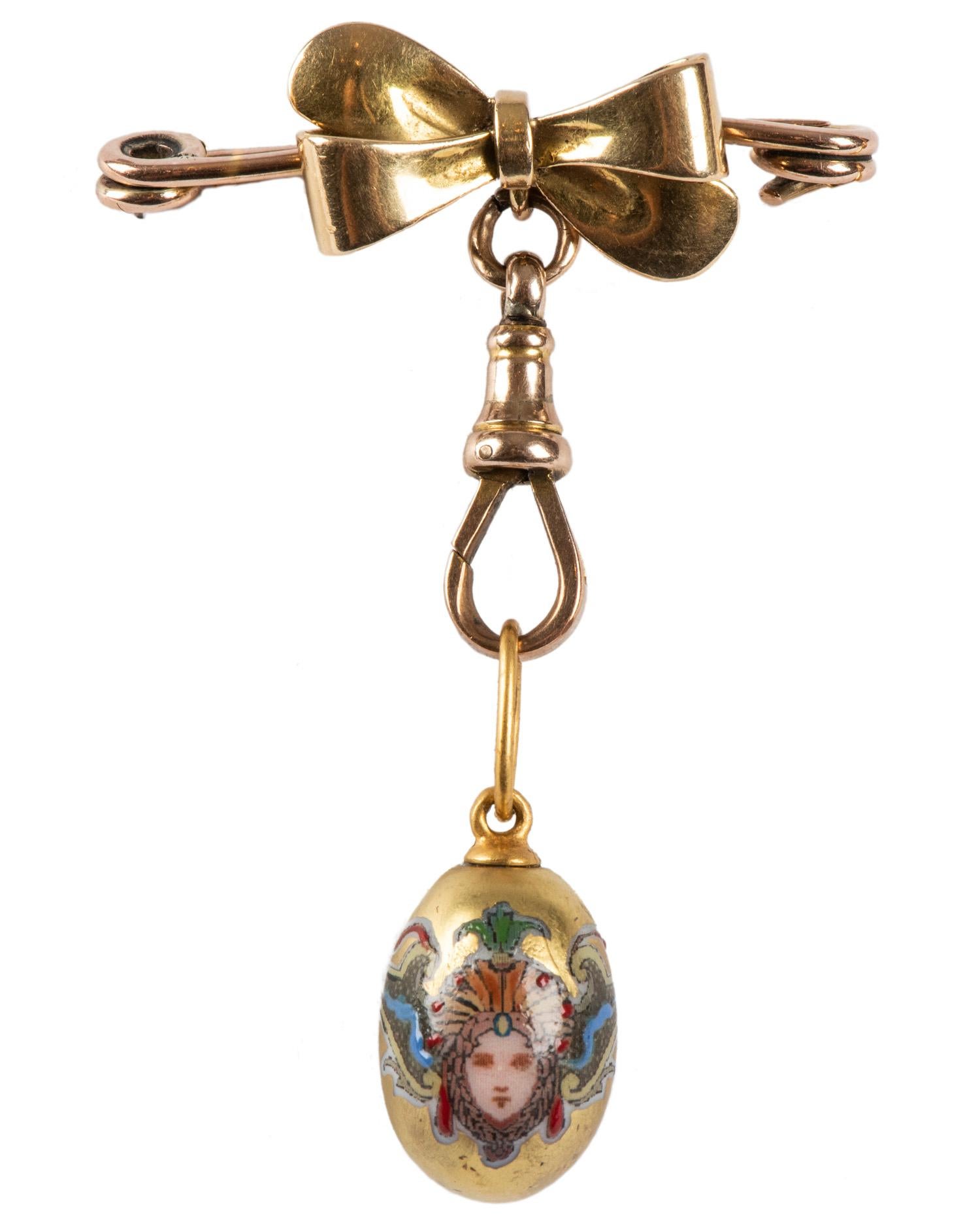 A Victorian pin designed as an 18k yellow gold bow on a 14k rose gold pin, suspending a Russian hand-painted porcelain gilded egg pendant, inscribed XB for Xristos Voskrese, a Russian Easter proclamation on pink ground..  

The pin circa 1890, the