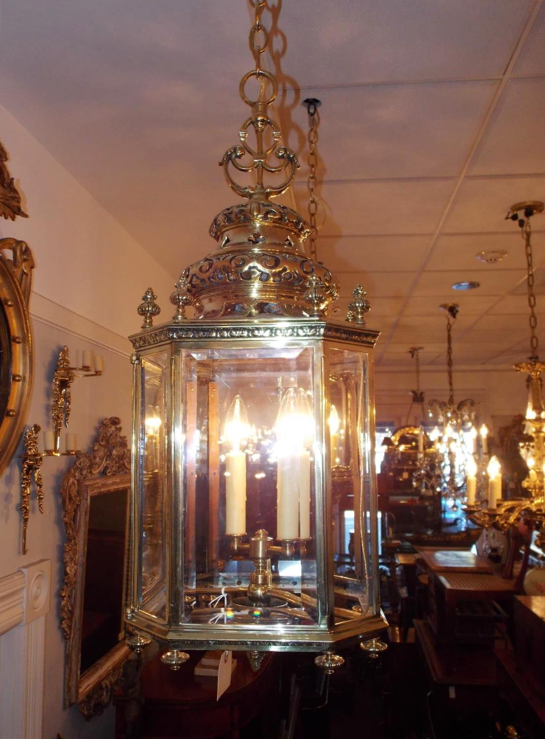 English brass octagon hanging hall lantern with a centered scrolled acanthus ring over a decorative bulbous two tiered hand chased pierced dome, original beveled glass with a single hinged locking door, eight upper floral finials with a filigree