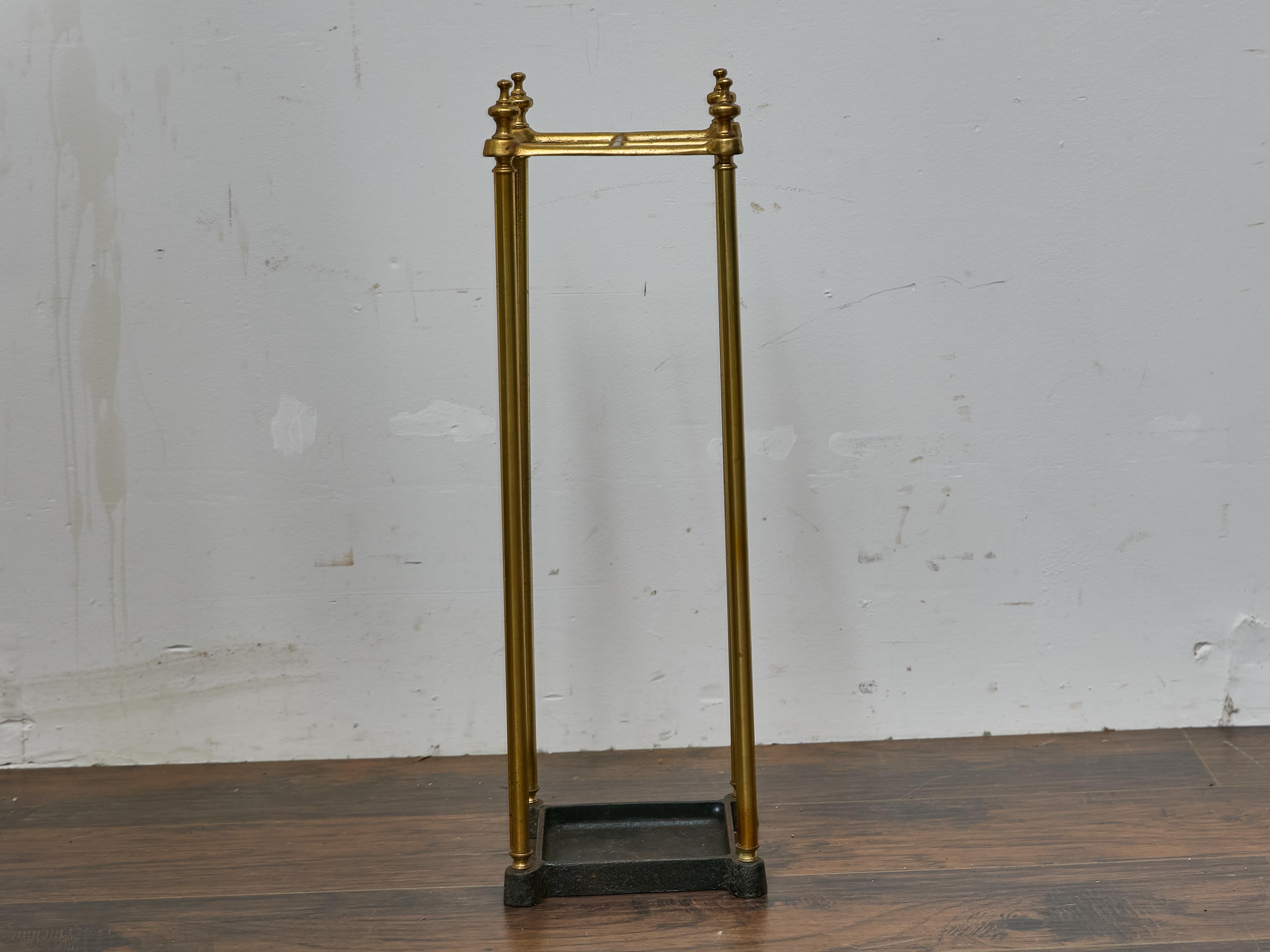 This early 20th-century English umbrella stand features a simple, well-balanced design executed in tubular brass with a square openwork top and a cast iron drip tray. The structure is supported by four slim brass rods that terminate in small feet
