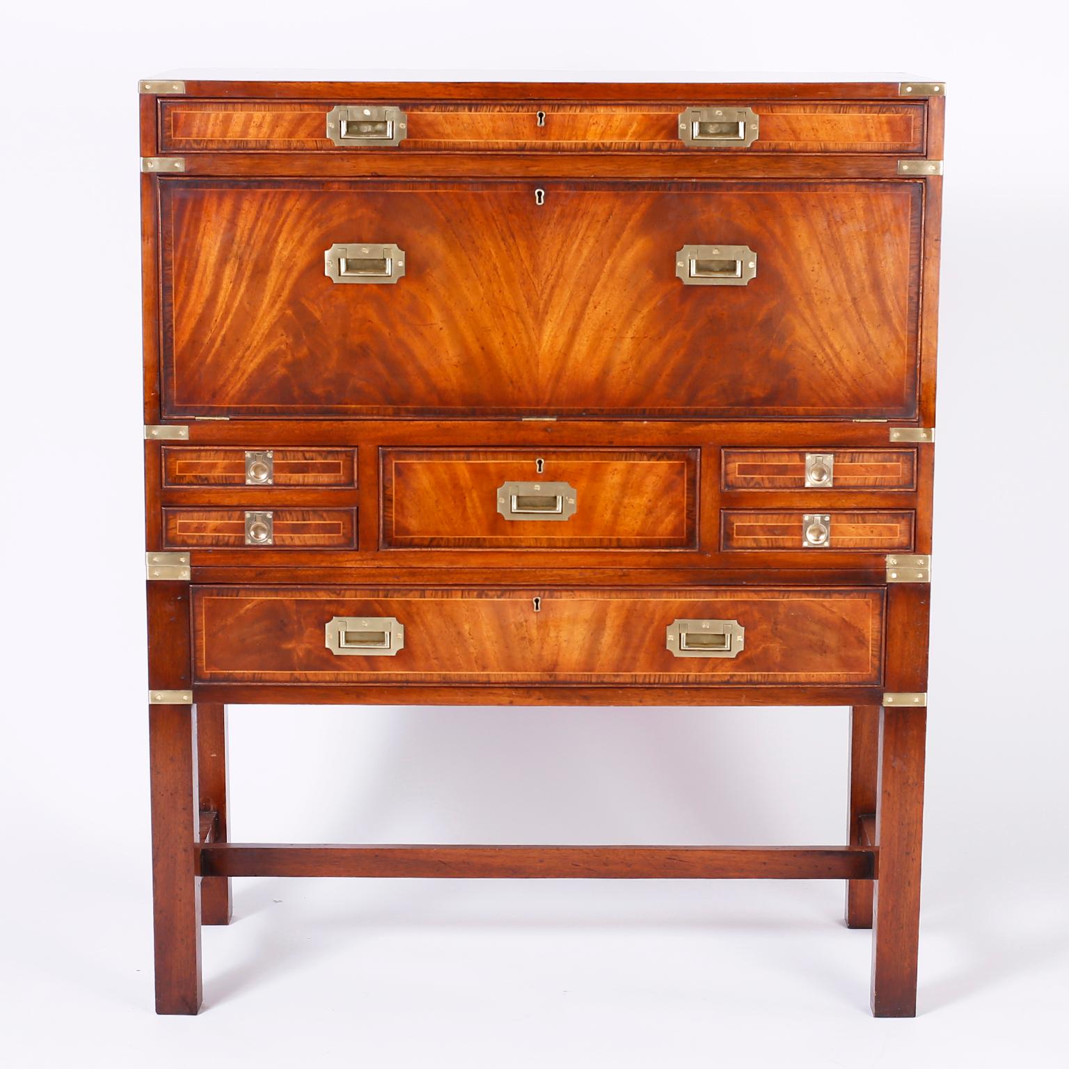 Antique English campaign chest on stand with seven drawers in assorted sizes, brass hardware and featuring exotic cuts of flame mahogany and a fall front butlers desk with slots, drawers and retaining the original tooled olive green leather.