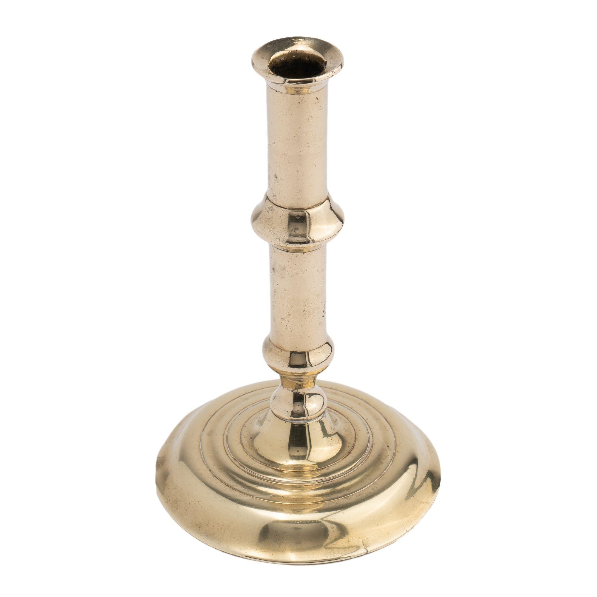 Cast brass cannon barrel candlestick with medial raised ring above a knob over waisted pedestal peened to a circular domed base.
Birmingham, England, circa 1720-40.