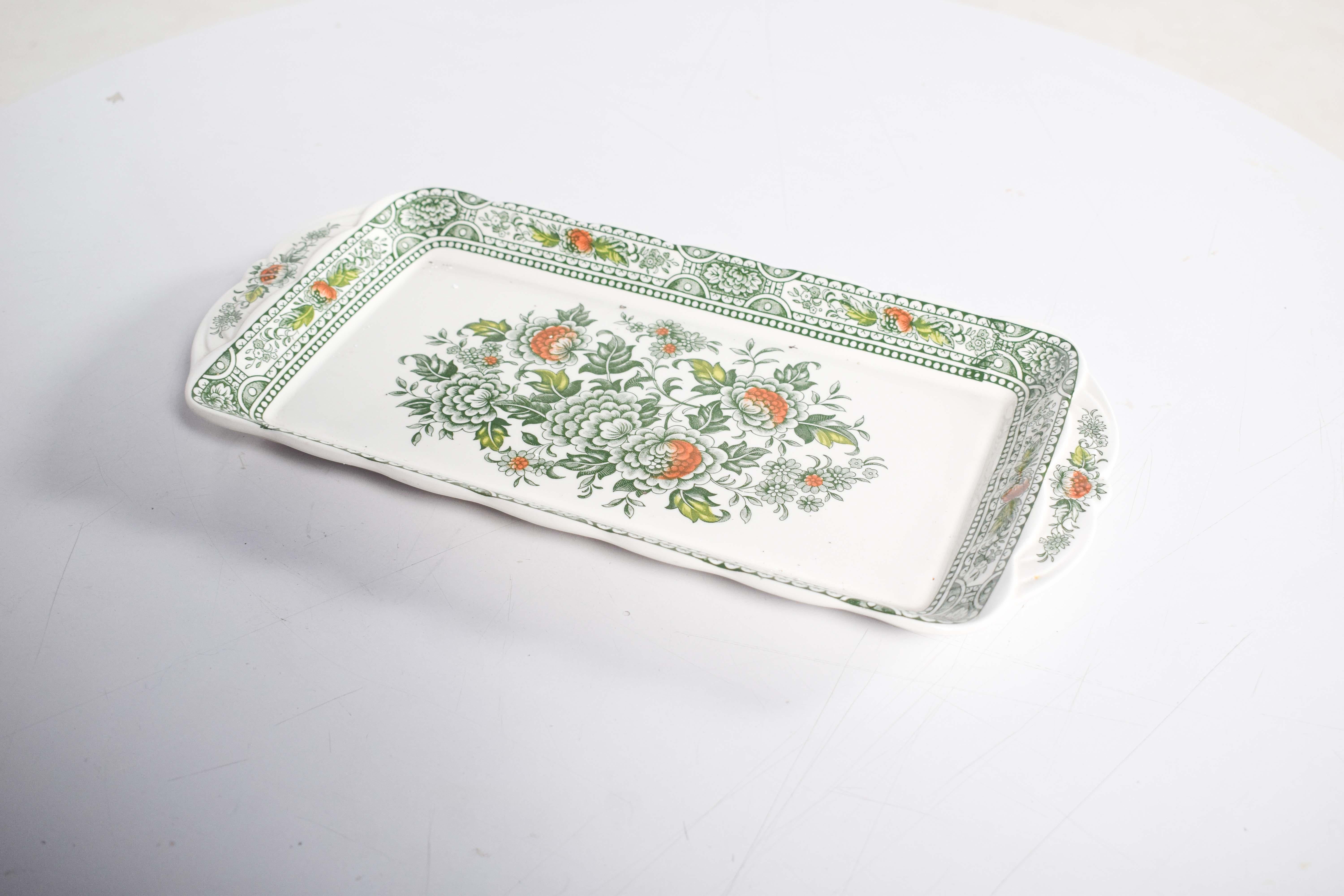 Esta elegante fuente de porcelana inglesa de Canterbury muestra un diseño floral clásico, reflejo de la fina artesanía de Staffordshire, Inglaterra. En su centro, un vibrante ramo de flores delicadamente detalladas se representa en varios tonos de