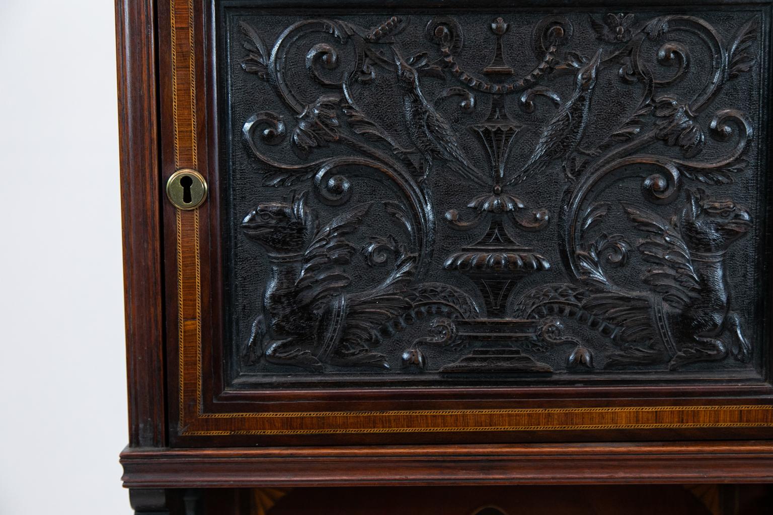 English carved inlaid cabinet is carved with birds and floral arabesques. The upper door is crossbanded with ebony, boxwood, and sandalwood inlay. The lower panel is inlaid with a classical urn and quarter fans. The top section is supported by