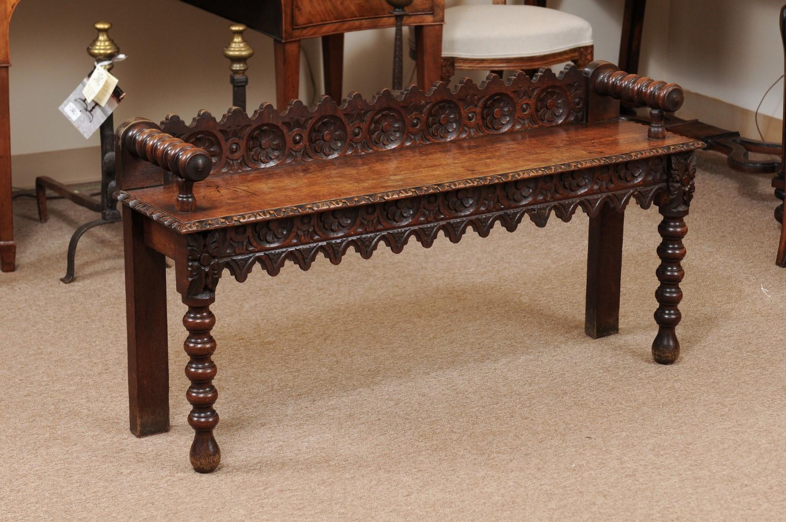 Late 19th century English oak hall bench featuring carved shaped back rail and apron with foliage carving and bobbin turned arms and front legs.
