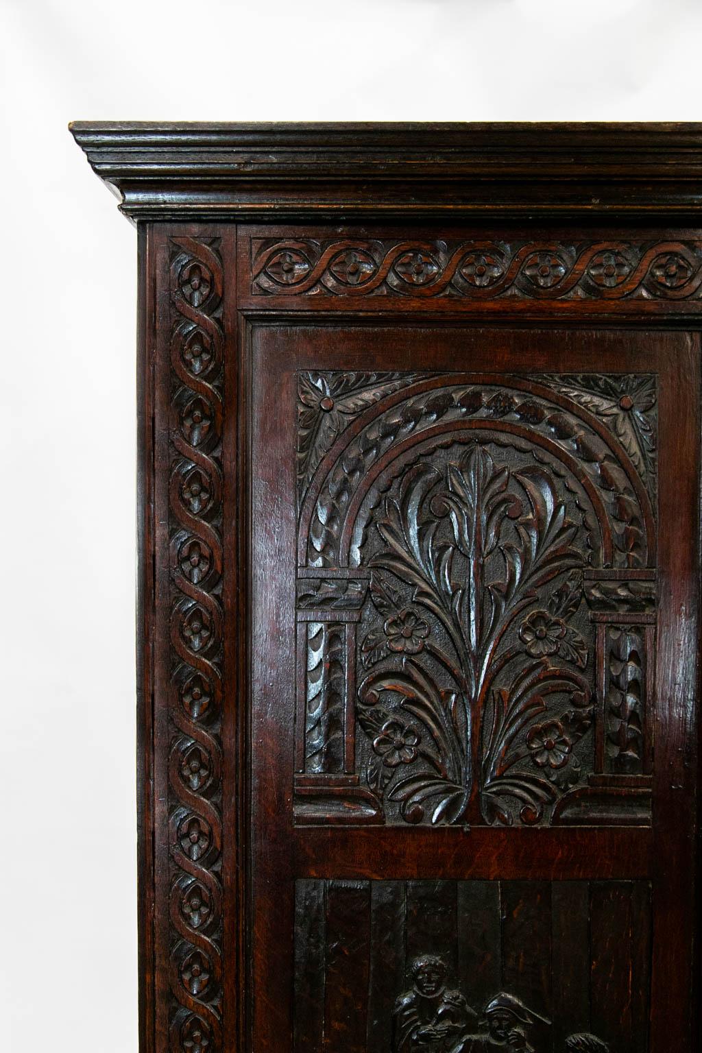 This oak cabinet on stand has two doors with recessed panels framed by repeating carved interlaced vines. Each door has two panels. The upper panel has a carved archway with a floral carved plant on a stipled background. The lower panels have male