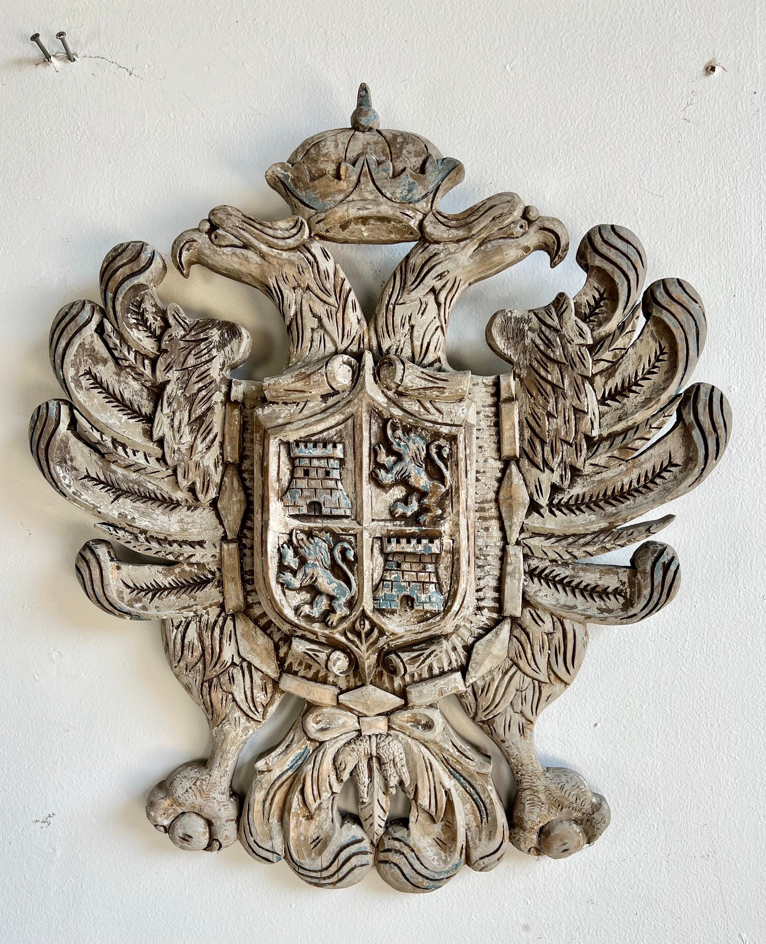 English Carved Shield w/ Coat of Arms C. 1930's. The shield depicts a pair of eagle heads with a crown. The shield also depicts a coat of arms with a pair of towers and a pair of lions. It is a great decorative piece for your home.