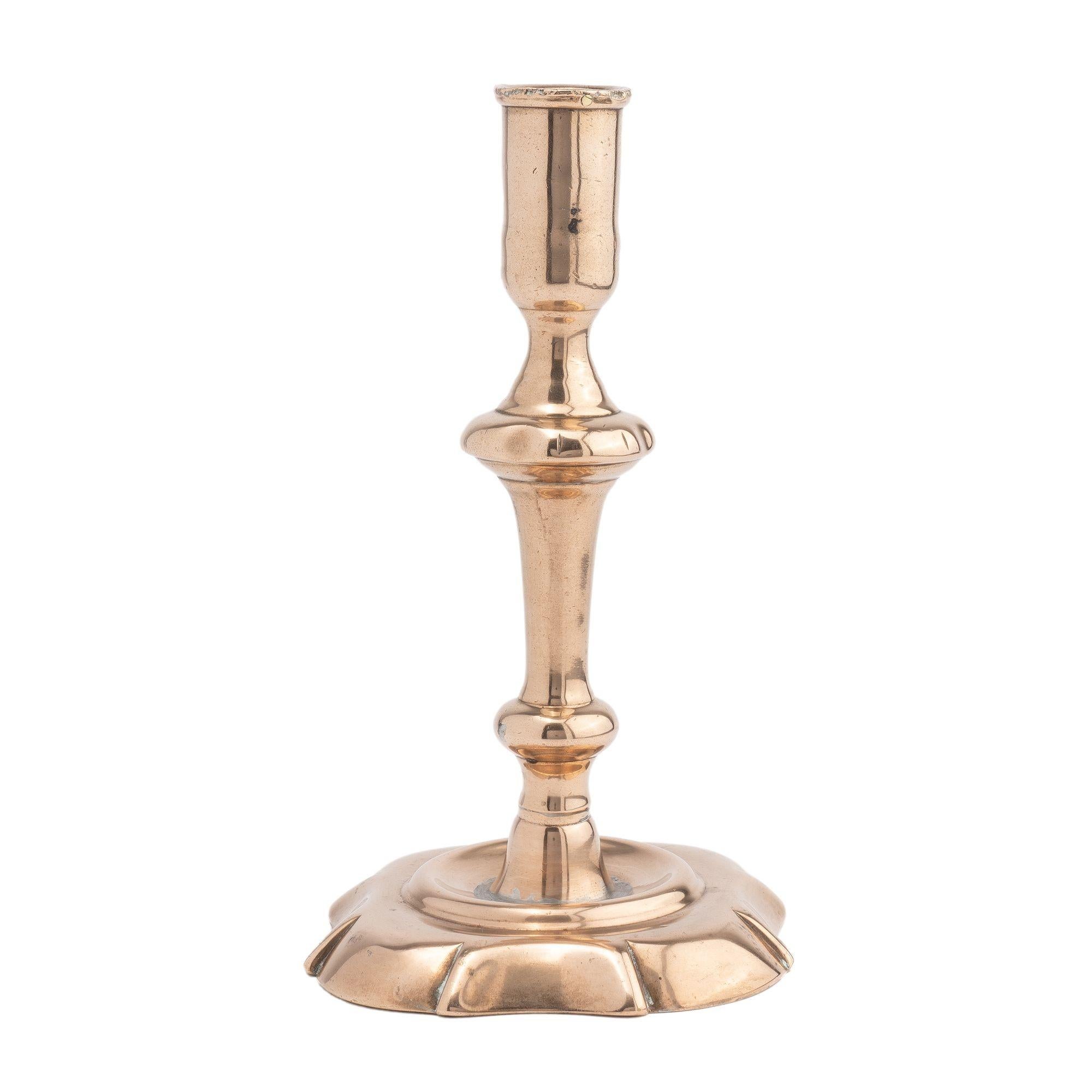 Hollow core cast bell metal brass Queen Anne candlestick. The candlestick features an elongated candle cup on a waisted turning to a lobed knob. The knob is supported by an inverted trumpet turning on a knob, waist, and corner lobe with side cut