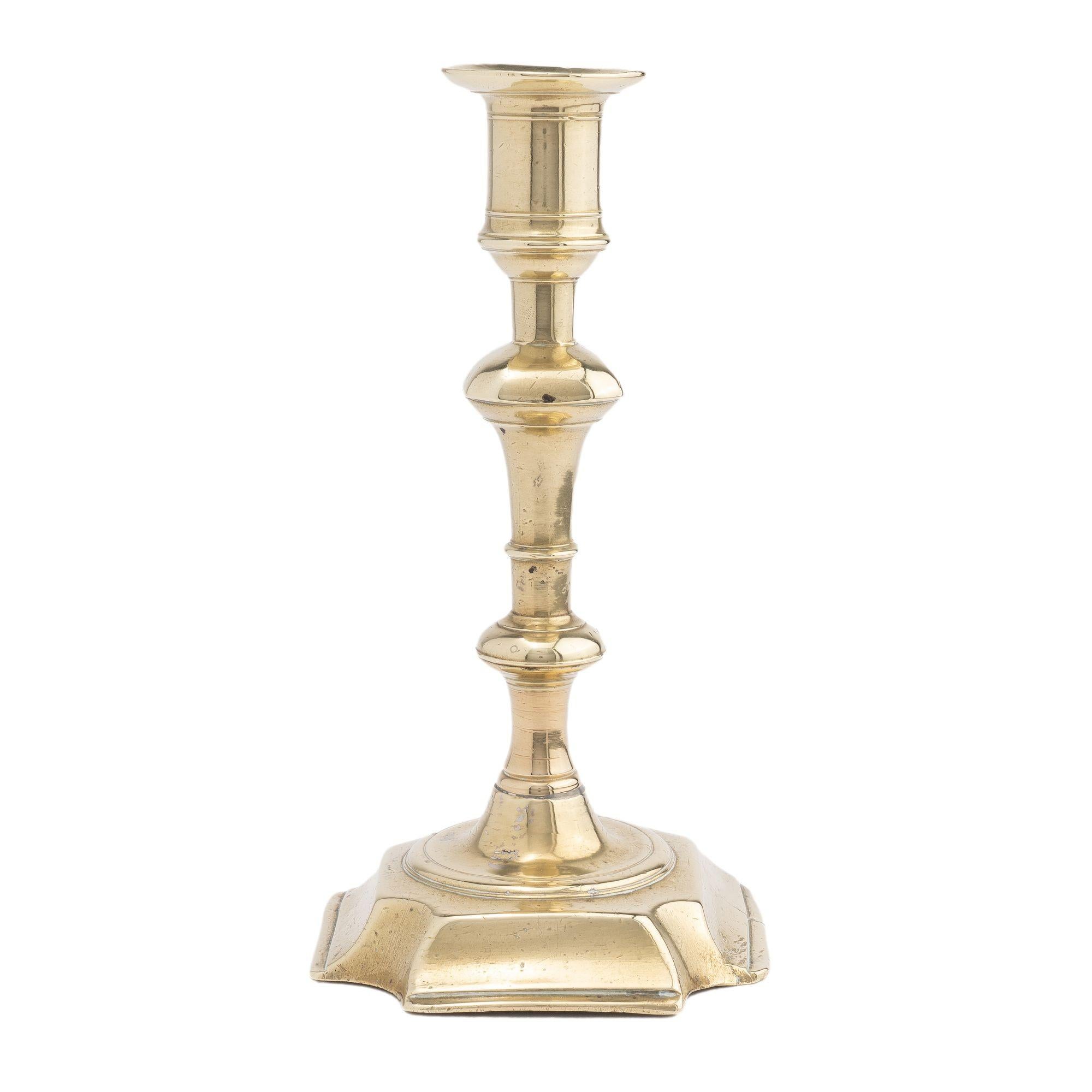 Cast brass Queen Anne candlestick with urn form candle cup with circular bobeshe, supported by a baluster with a four sided knob above ring and knob turnings peened to a raised square base with cove cut corners. The shaft has been cast in two parts