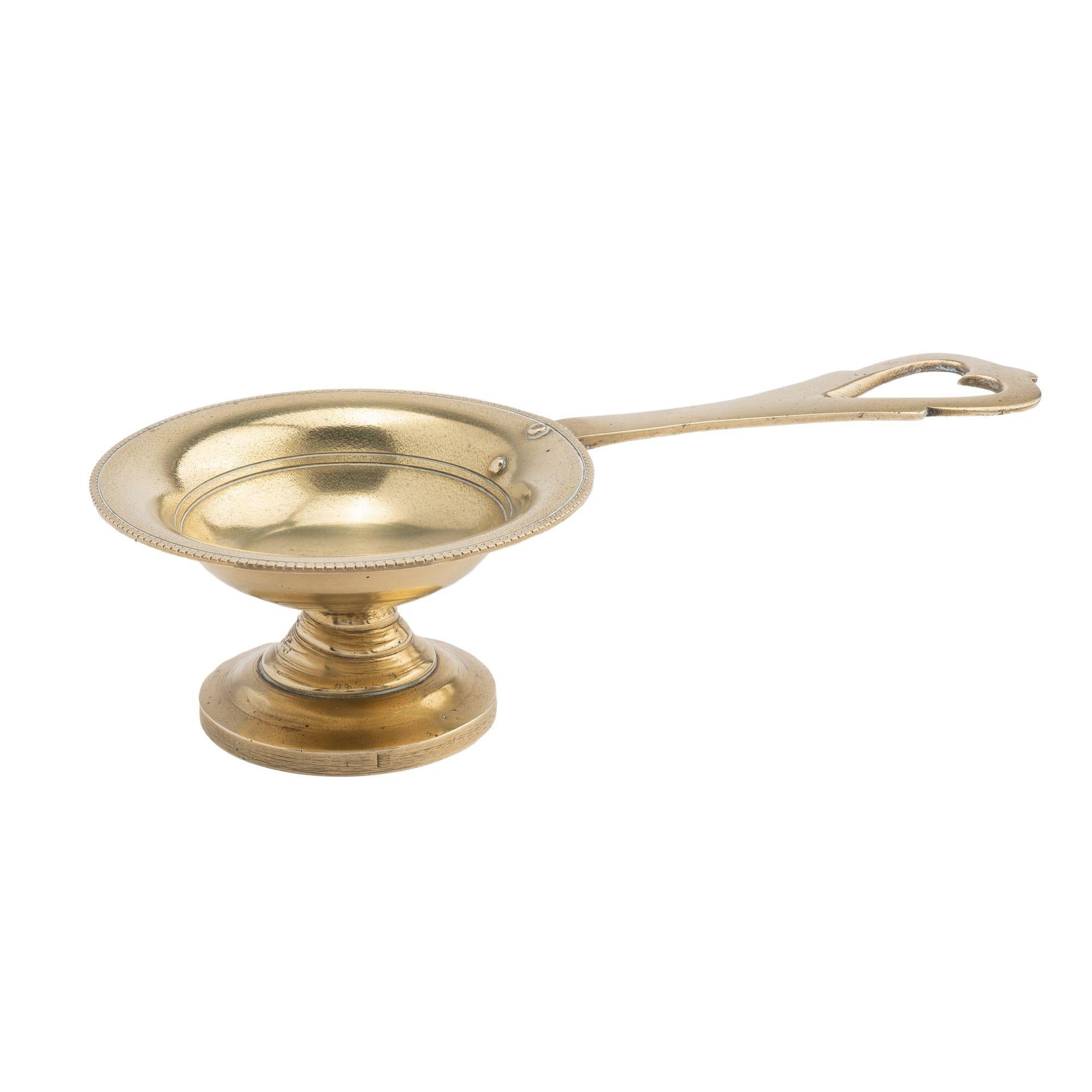 Shallow circular cast brass taster dish with beaded edge on a circular pedestal base. The flaring rim is rivet mounted with a long handle pierced with a heart shape at the terminal.
English, fourth quarter 1700’s.