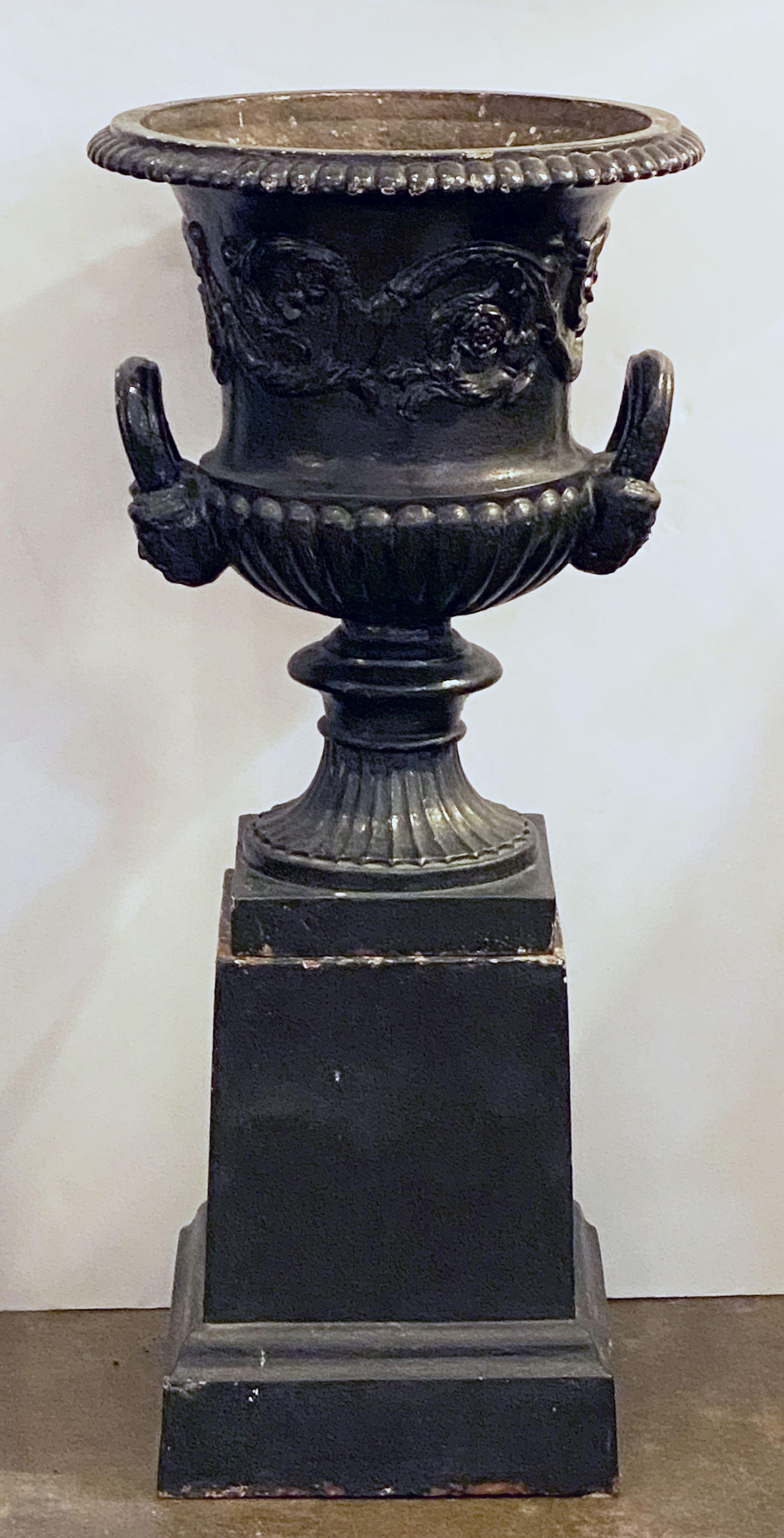A fine pair of English cast iron urns (or garden planter pots) on plinths from the Regency Era, circa 1820s - each urn featuring a classical style design with everted dentil rim, quarter-lobed body with foliate relief, and flared masked handles on
