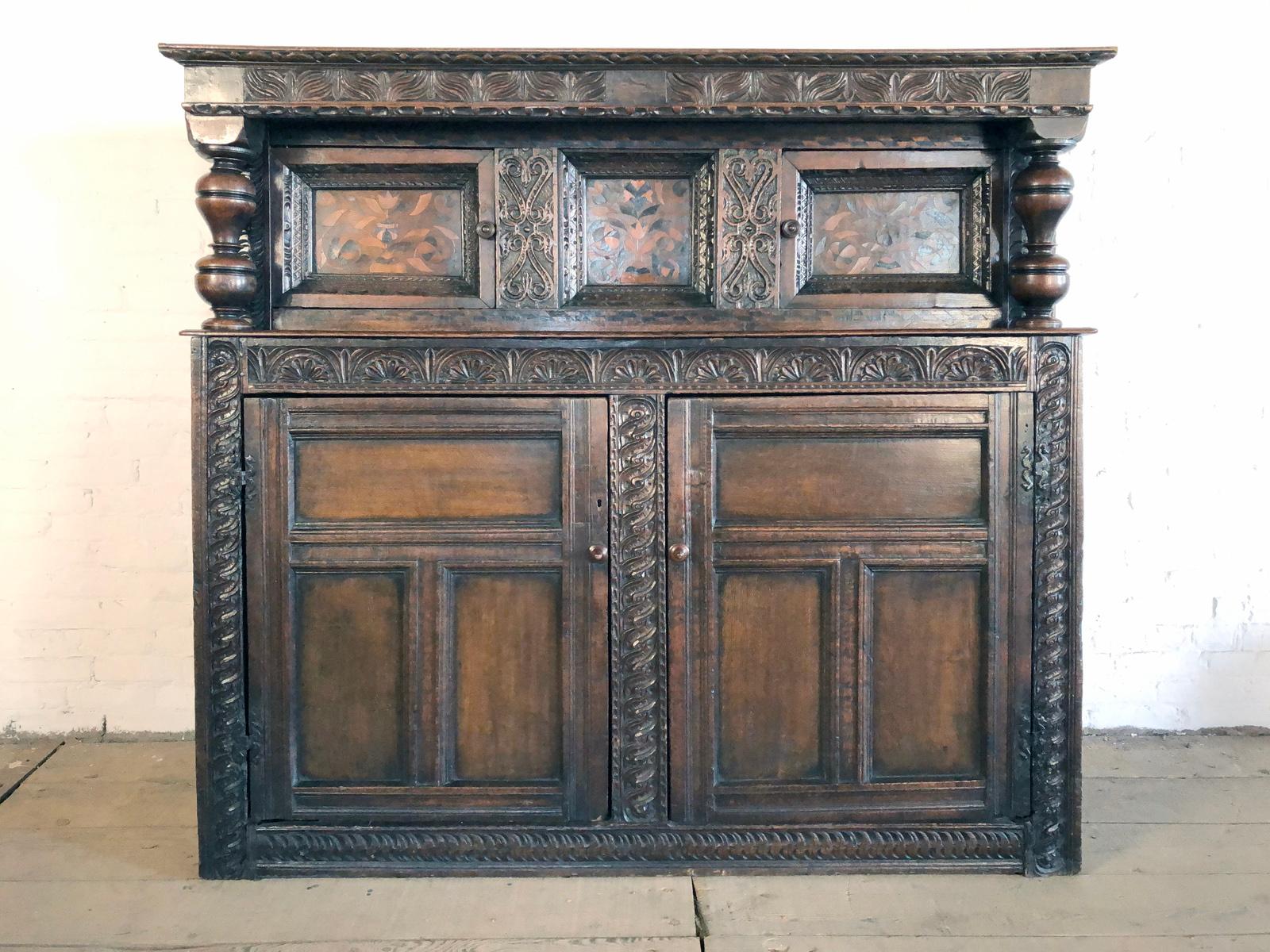 Large English Charles I Oak and Inlaid Court Cupboard, Yorkshire circa 1640, having a boarded top with a delicately carved edge above a leave-carved frieze, the upper part with turned baluster supports on each end, flanking two doors with deeply