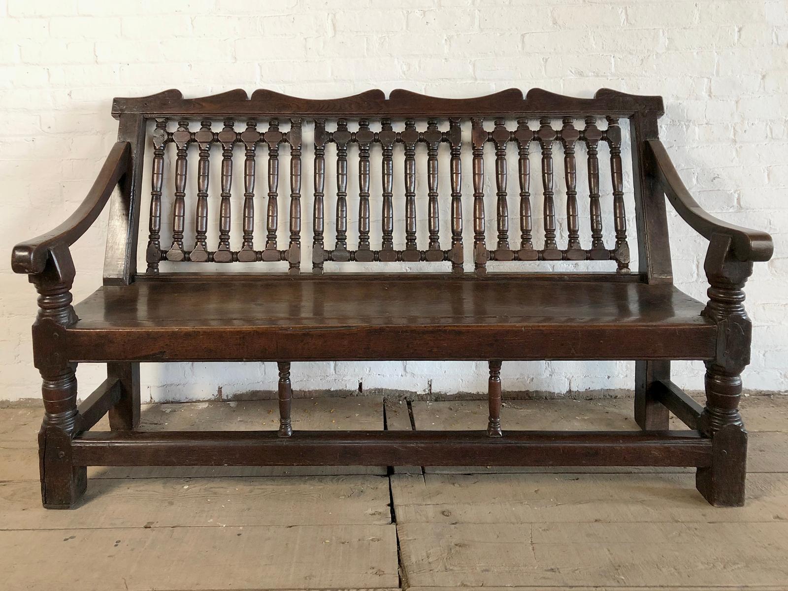 A very rare and unique English Oak bench, of triple chair-back form with three sections of turned spindles, topped by a wavy carved cresting, downswept open arms on short turned front supports, the beautifully figured plank seat supported by turned