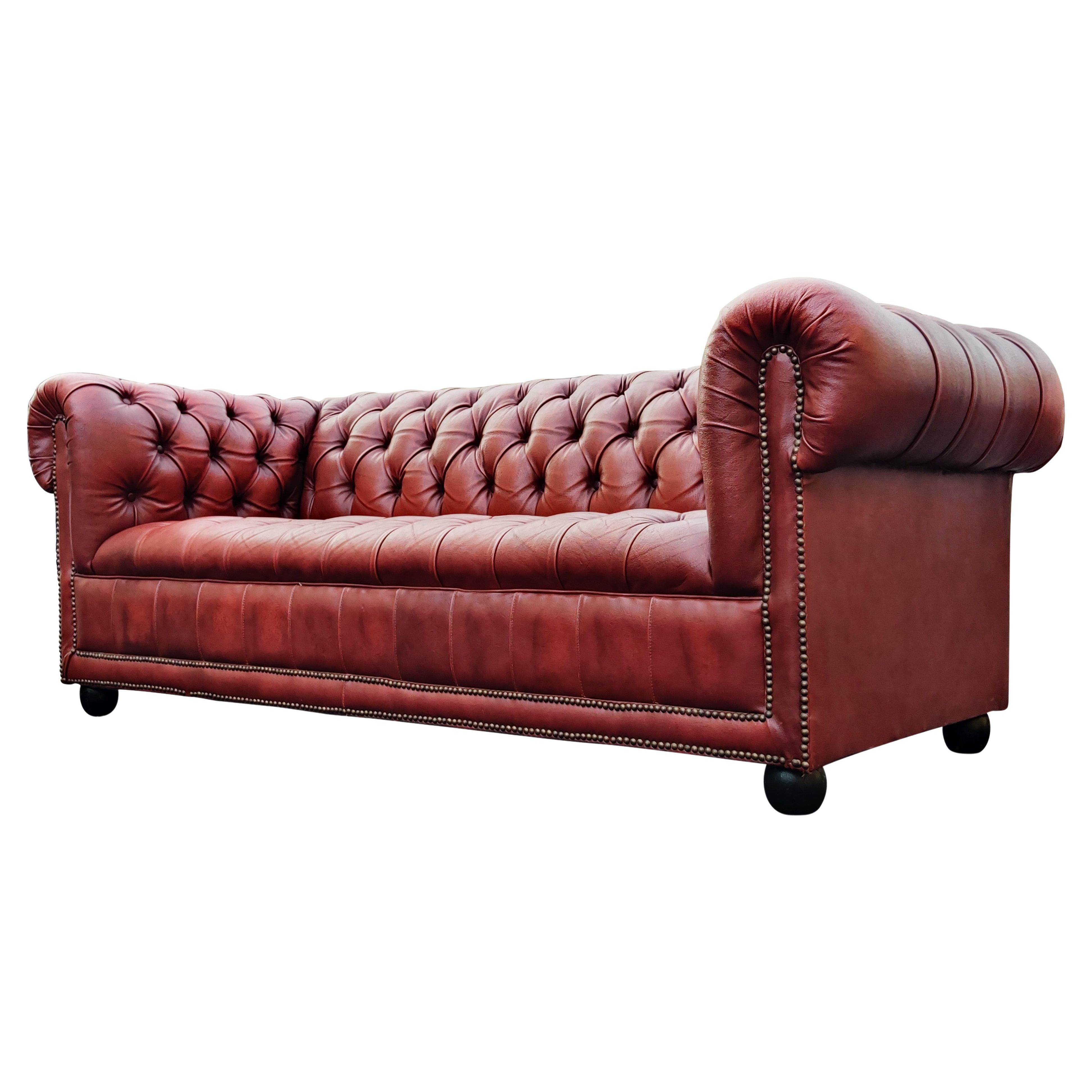 A classic English Chesterfield sofa or settee featuring an oxblood or cordovan leather. The frame is crafter with large English scrolled arms, having a tufted leather seat, arms, and back. Brass tacks or nail-heads accent the hand-tailoring