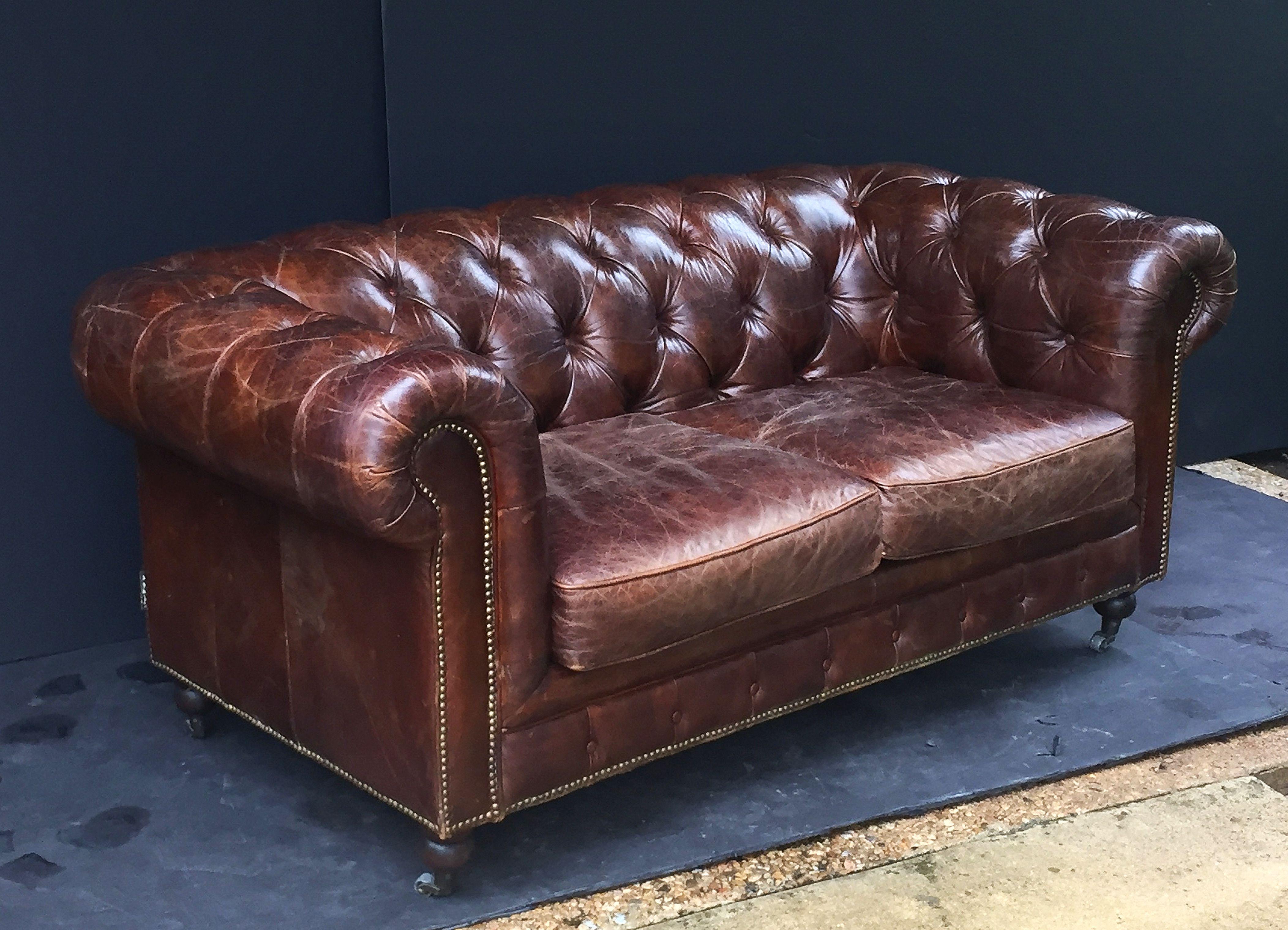 A fine, comfortable English Chesterfield sofa in vintage brown or tobacco leather featuring button-tufted back and arms, two soft leather fitted seat cushions, and beaded-nail trim design, resting on turned feet with casters.

The Chesterfield