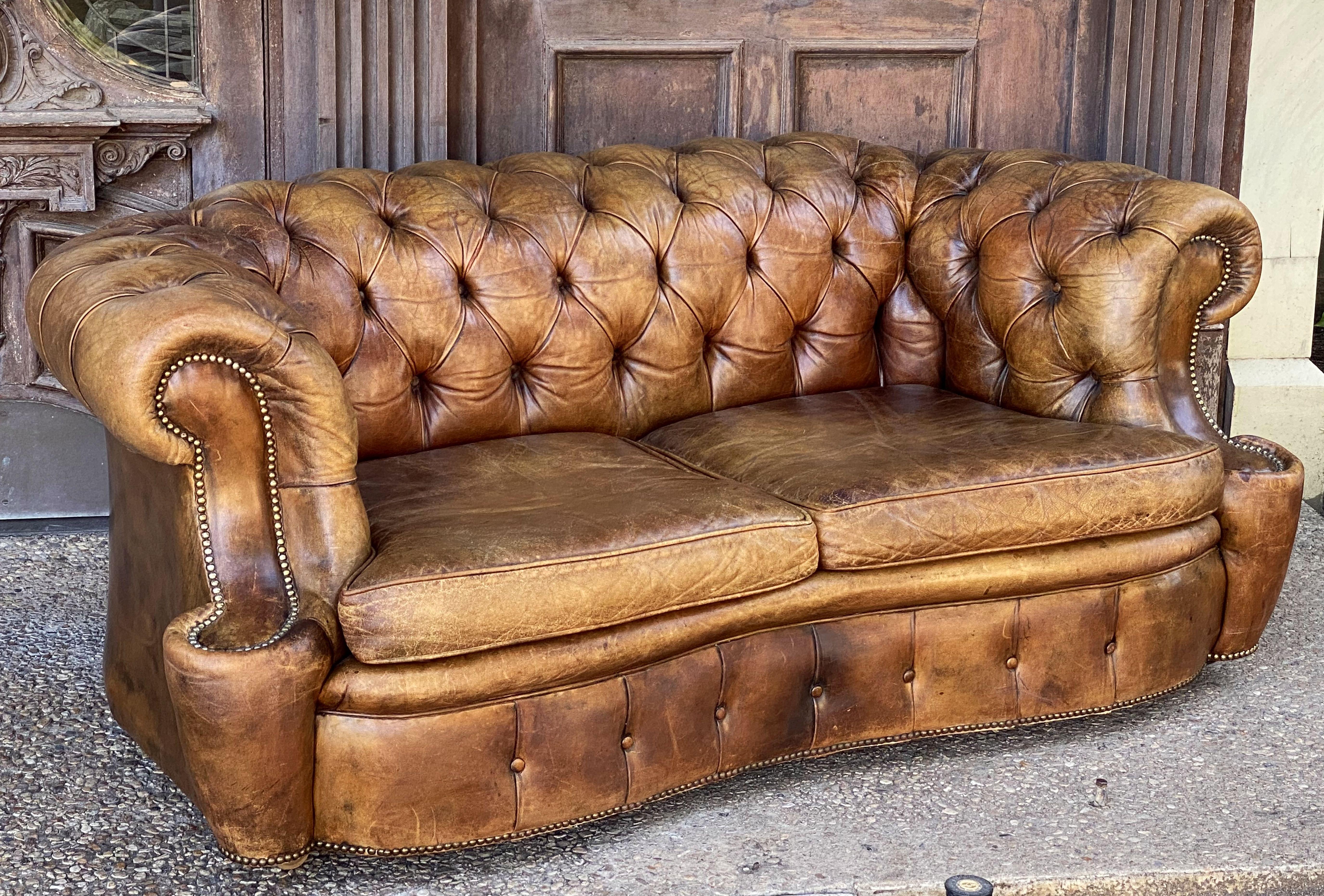 A fine, comfortable English Chesterfield sofa in vintage leather featuring a serpentine profile with button-tufted back and arms, two soft leather fitted seat cushions, and beaded-nail trim design, resting on removable metal casters.

The