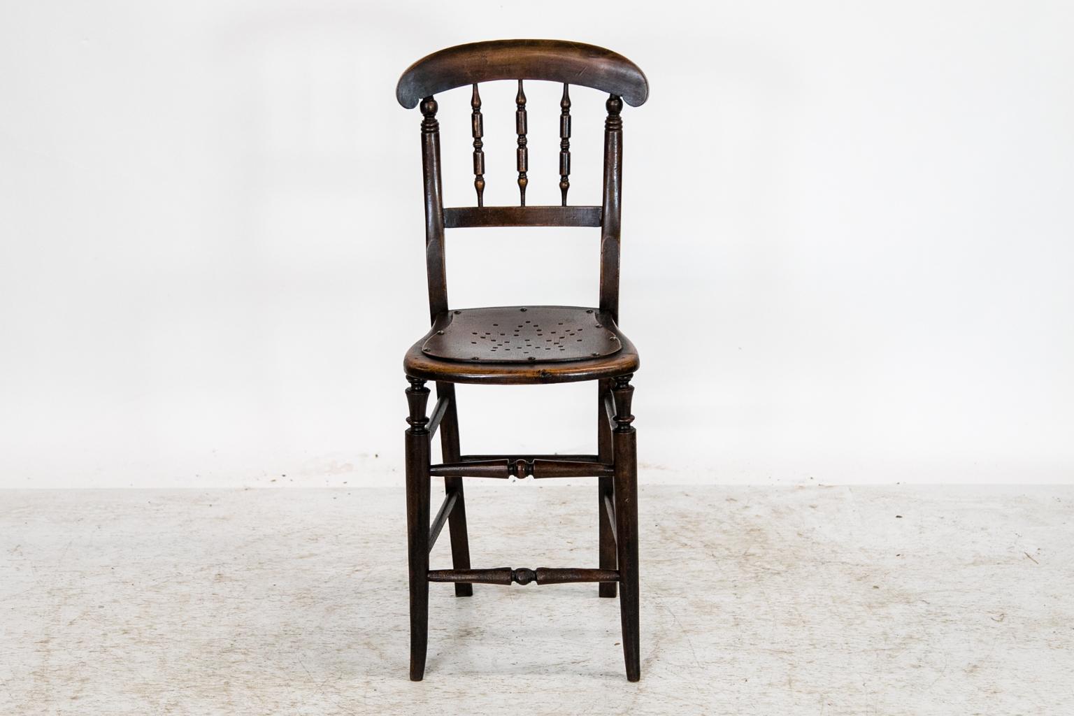 This child's chair has a scrolled crest rail above three turned spindles in the back. The seat is the original pressed wood with a perforated star pattern. The front legs are turned with double turned cross stretchers and terminate with a slight