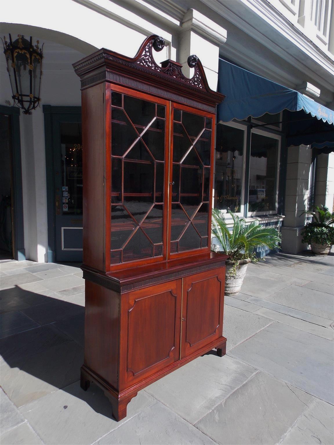 English Chippendale mahogany bookcase with a broken arched pediment, centered floral medallions, intricately carved fret work, dental molding, fluted gouge work, upper case with two hinged glass doors revealing four adjustable interior shelves,