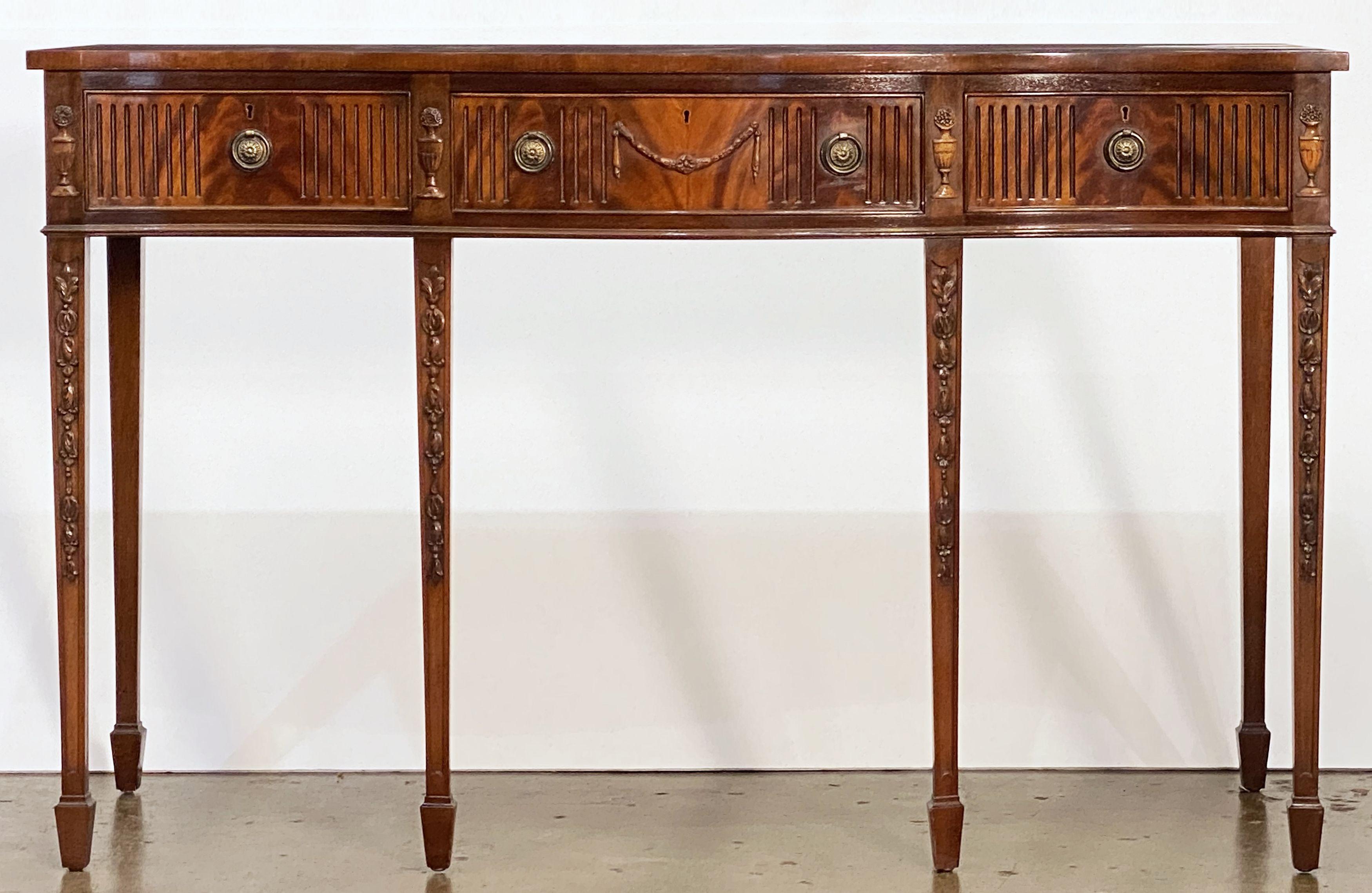 A large English console server or buffet table of mahogany in the Sheraton style, featuring a serpentine top over a frieze of three drawers, with beautifully carved urns in between the drawer fronts and down the tapered legs. 
The drawers of solid