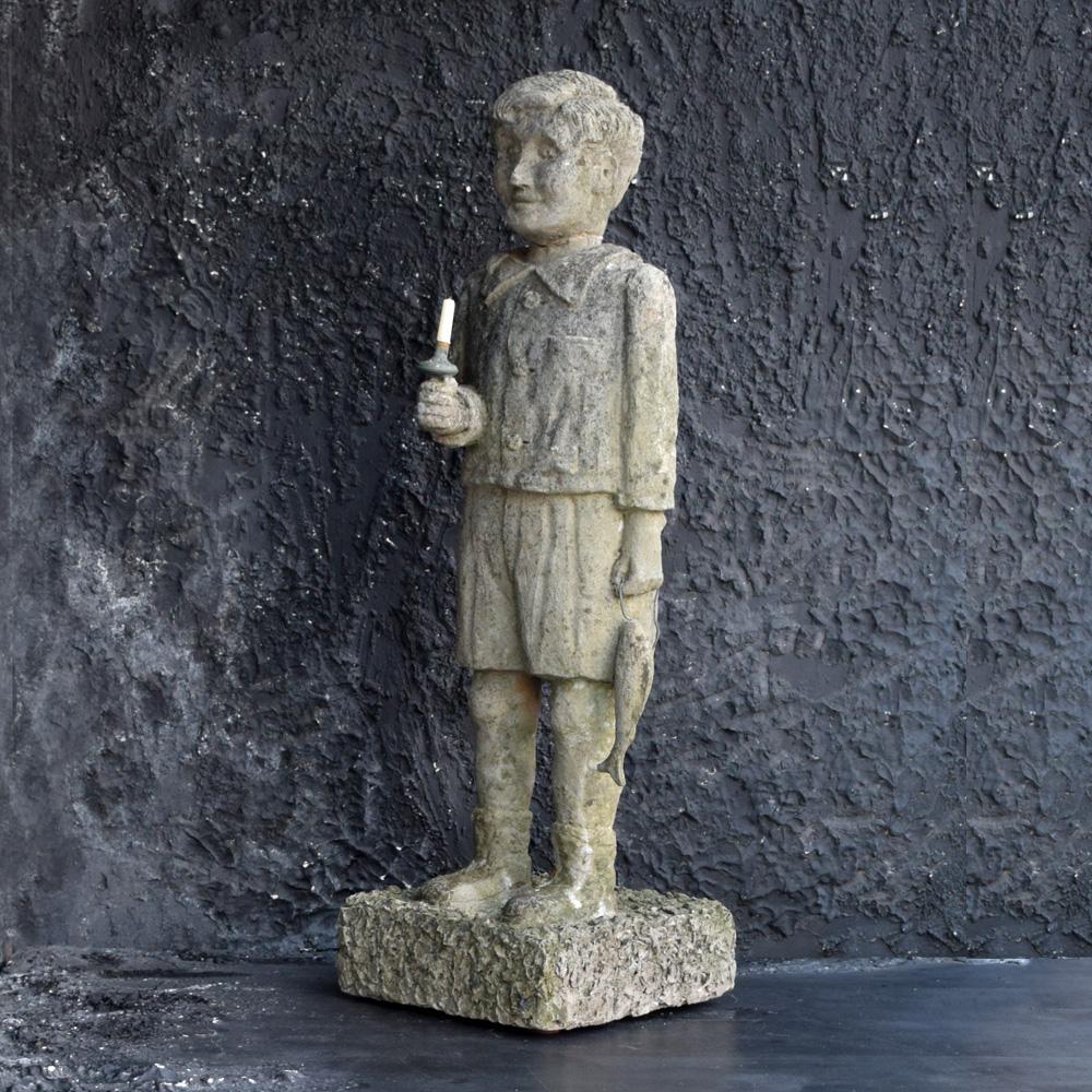 A mid-20th century stone statue of a young boy holding a fish and copper pole. This statue can easily hold a candle as displayed in the photos provided and could easily be placed as an internal Folk Art centrepiece. This stone figure has a very
