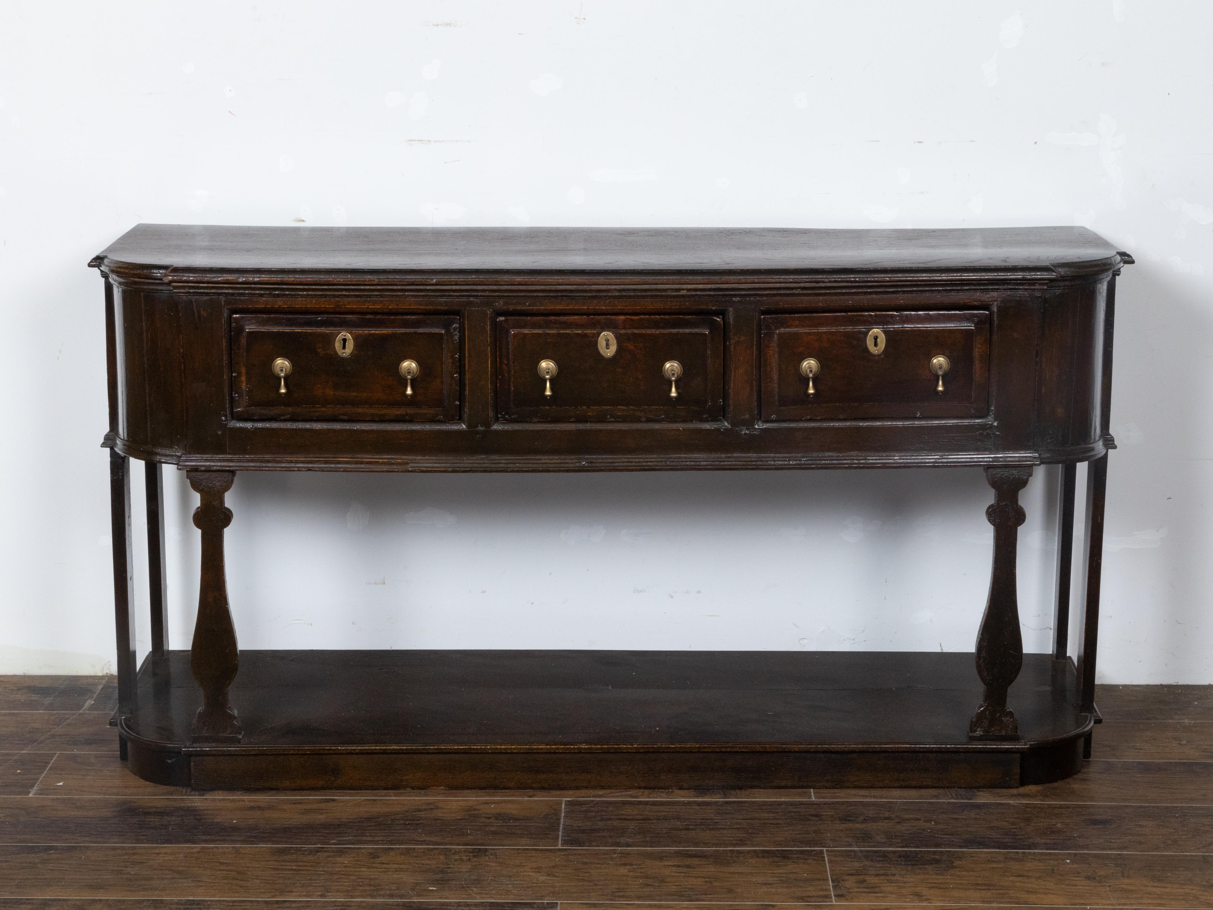 An English Georgian period oak dresser base circa 1800 with three drawers, carved legs, brass hardware including teardrop pulls, pot holder shelf and dark patina. This English Georgian period oak dresser base, circa 1800, exudes a sense of