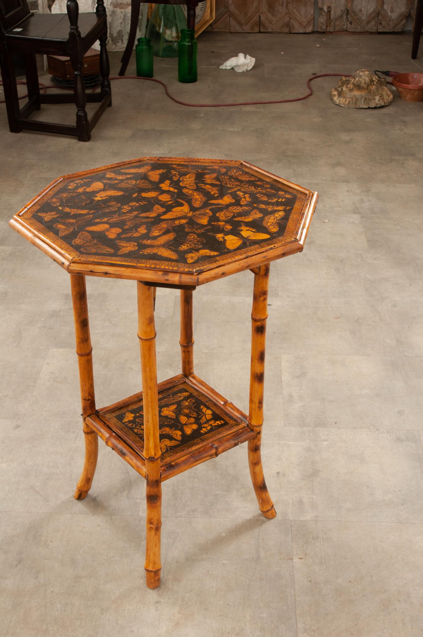 A chic English decoupage bamboo table in the Aesthetic Movement style of the late 19th century. This piece features a speckled bamboo frame supporting two tiers of beautifully decoupaged shelves showcasing gilded butterflies and moths surrounded by