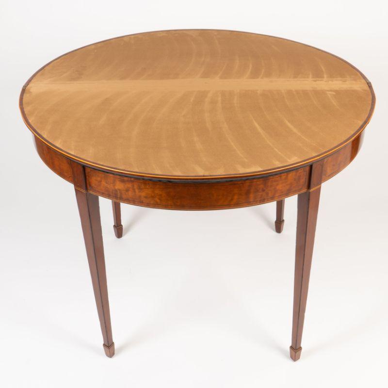 Circular flip top game table of mahogany solids and curly mahogany veneers with ebony edging and holly string. The table rests on a conforming demi lune apron interrupted by four legs dies which continue into four square tapered legs with holly