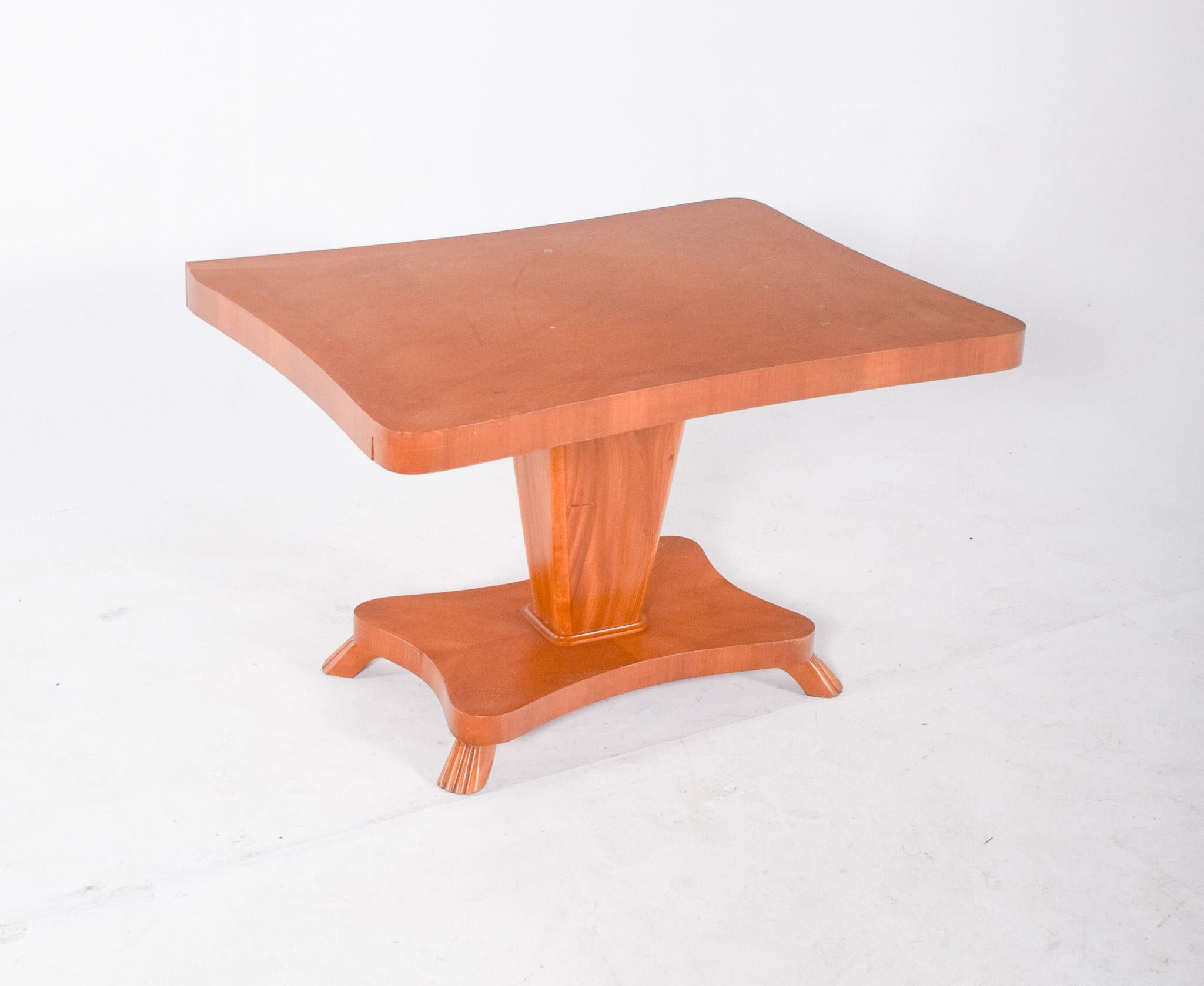 Dining table with centre leg. The top has a rectangular shape with curved edges and a light varnished finish. The base is made up of a wide column that connects to a square lower base also made of wood, with soft contours and four small, curved