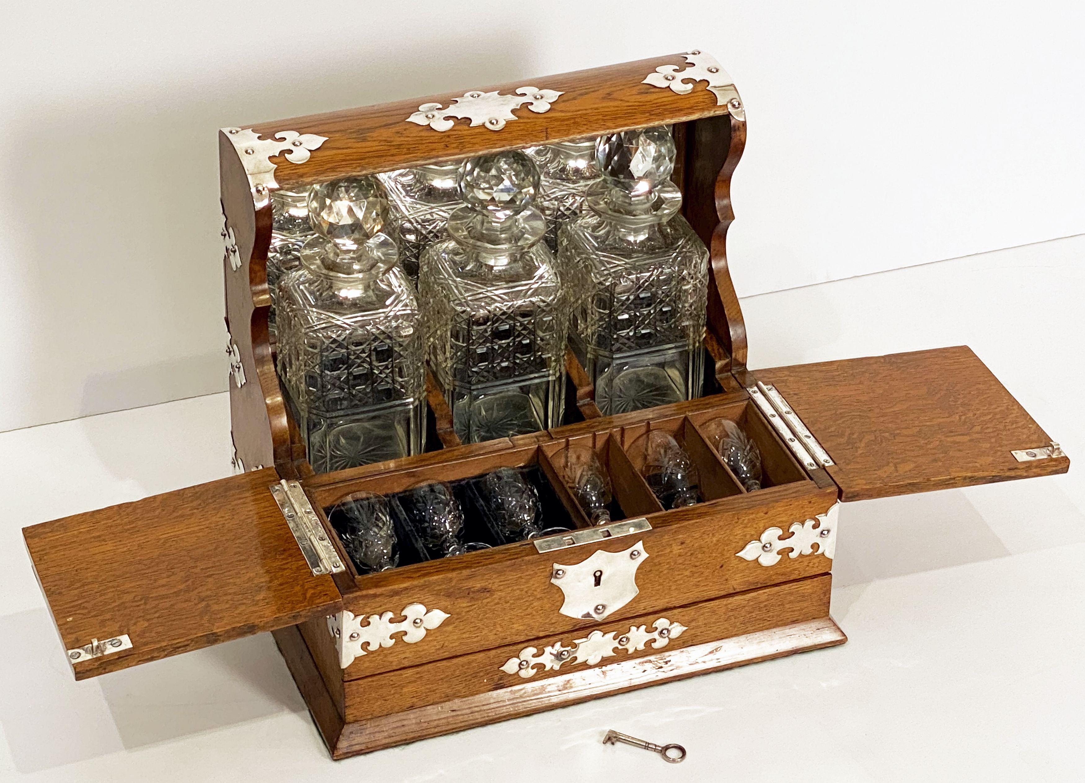 A fine English drinks tantalus of silver-mounted patinated oak with three decanters and a games drawer compendium from the Edwardian era.
Featuring a top mount enclosing three square cut-glass spirits decanters within a fitted mirror-back