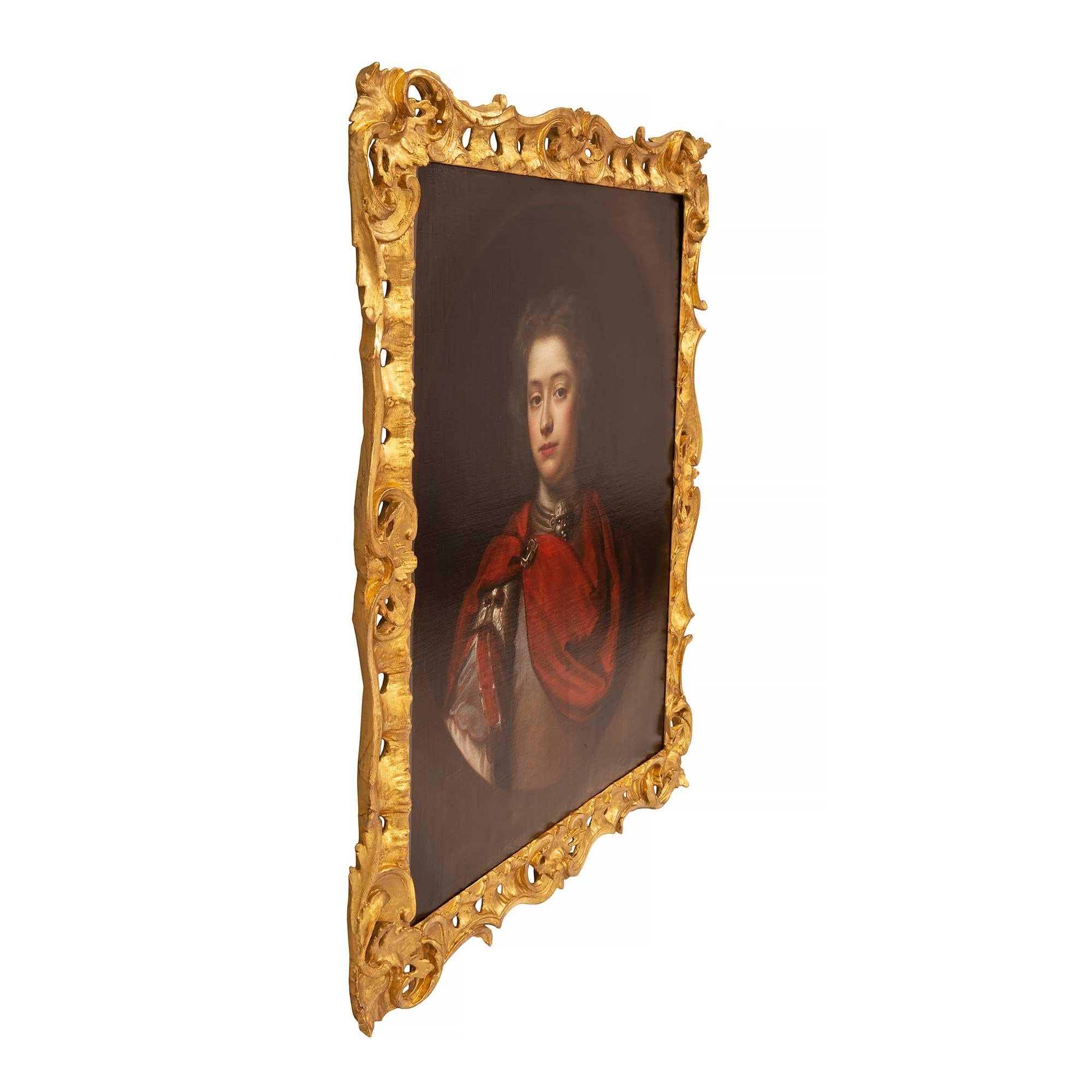 A lovely English early 19th century oil on canvas within a fine giltwood frame. The painting depicts a young nobleman in armor and draped in a red cape. He wears a pauldron and a fine necklace. Framed within an elegant foliate pierced giltwood