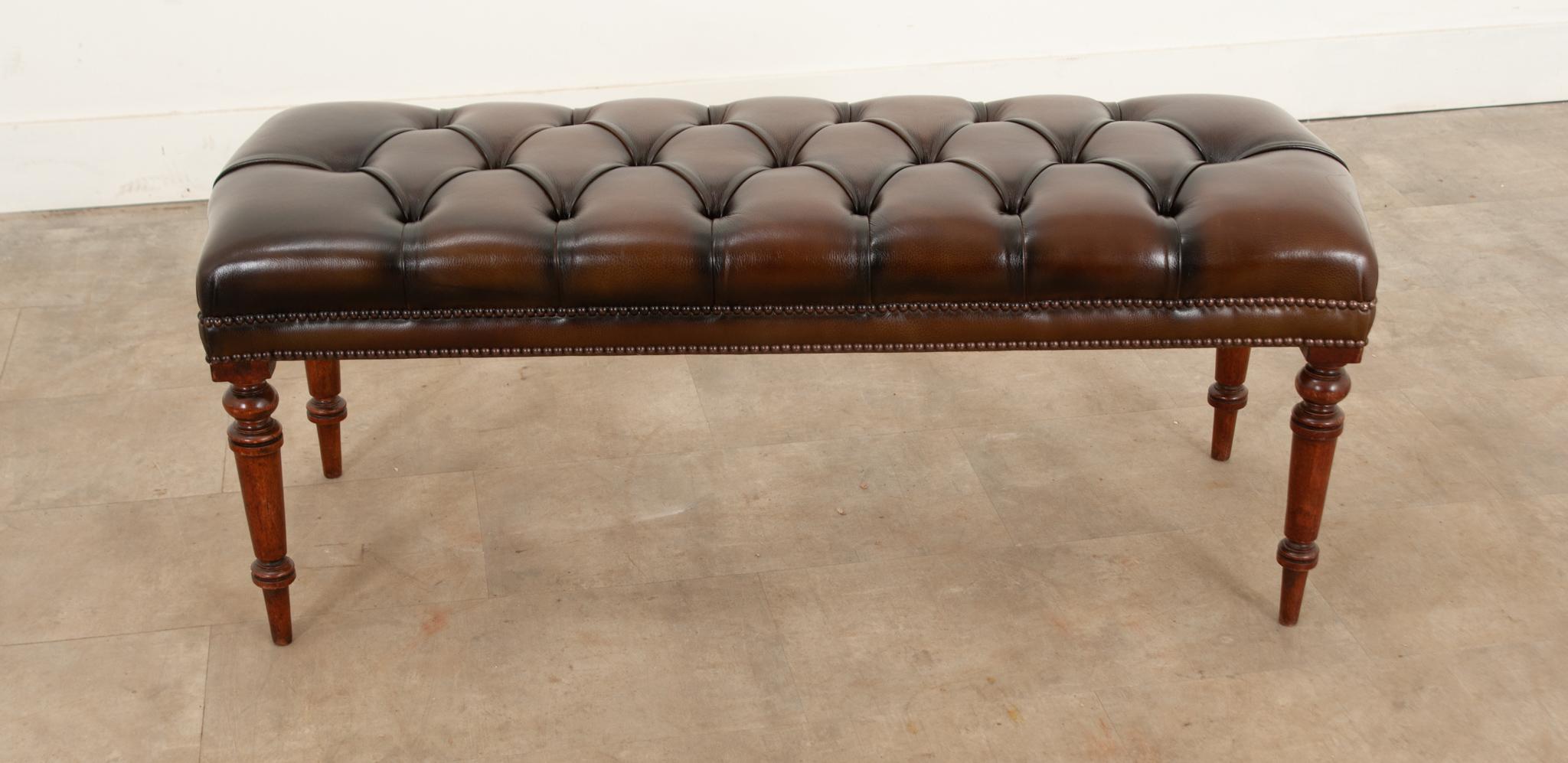 A handsome mahogany bench with beautifully turned legs, newly upholstered in tufted leather. The leather is a rich robust brown tone that gleams, with decorative rivets at the legs. For use as a comfortable seat or as a posh coffee table with use of