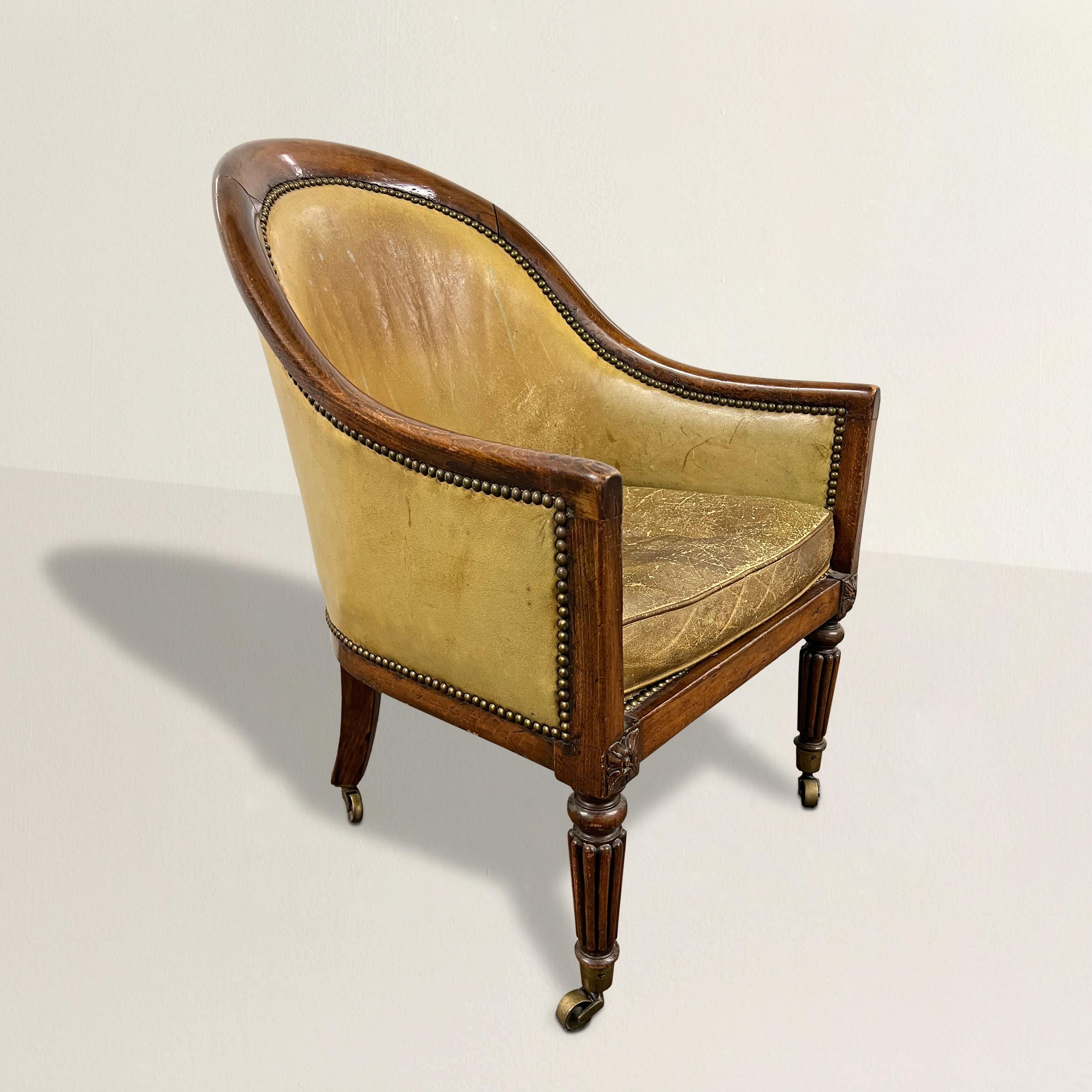 An inviting and enveloping early 19th century Early Victorian round back armchair with a wonderfully curved wooden frame, leather upholstery with a tight back and loose seat cushion, and standing on tapered fluted legs with brass casters. The