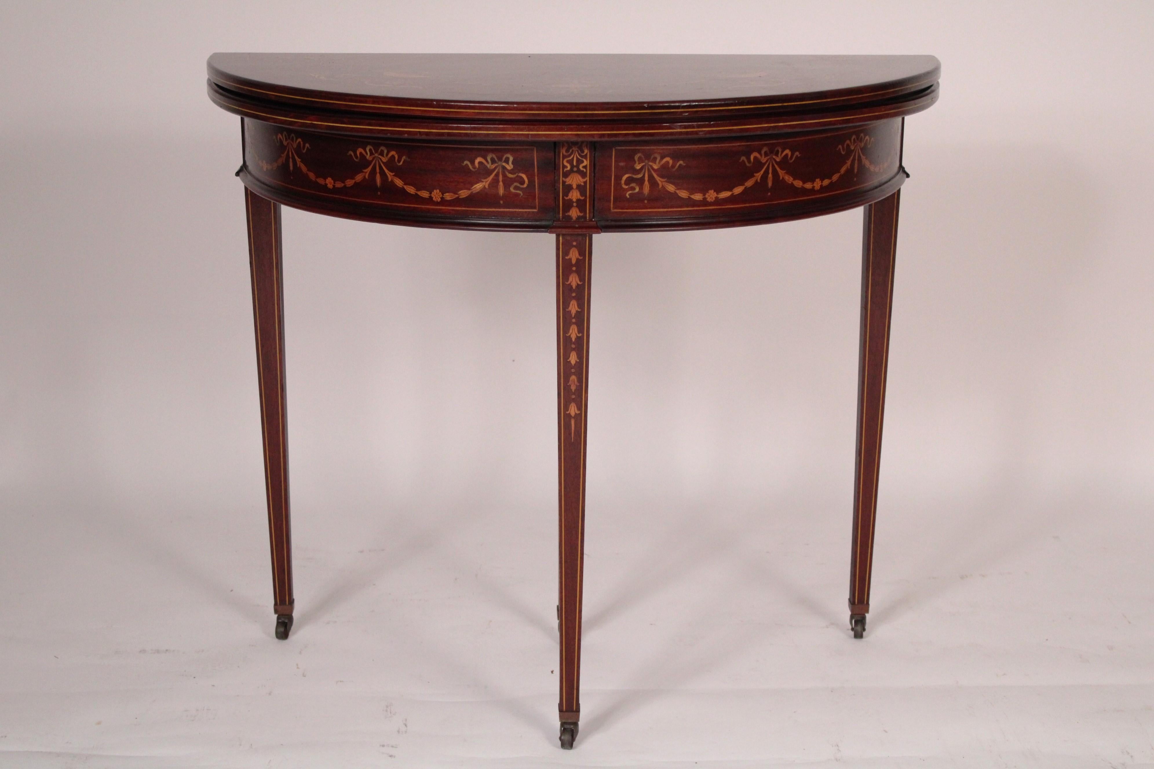 English Edwardian mahogany inlaid demi lune games table, circa 1920.
With a demi lune shaped top with swag and urn inlay, a frieze with swag and ribbon inlay the square tapered legs inlaid with bellflowers and string inlay, resting on brass casters.