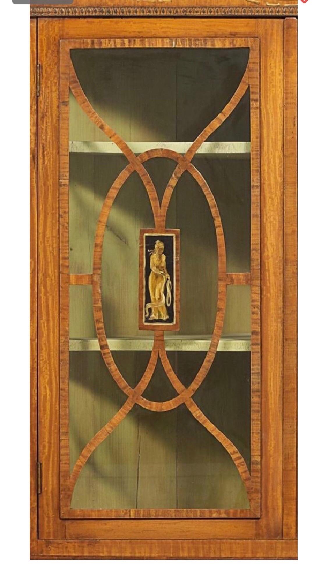 English satinwood and paint decorated corner cupboard from the Edwardian period. 2 part construction with neoclassical motifs painted throughout. Glazed doors on top and blind doors on the bottom- all resting on flared bracket feet.