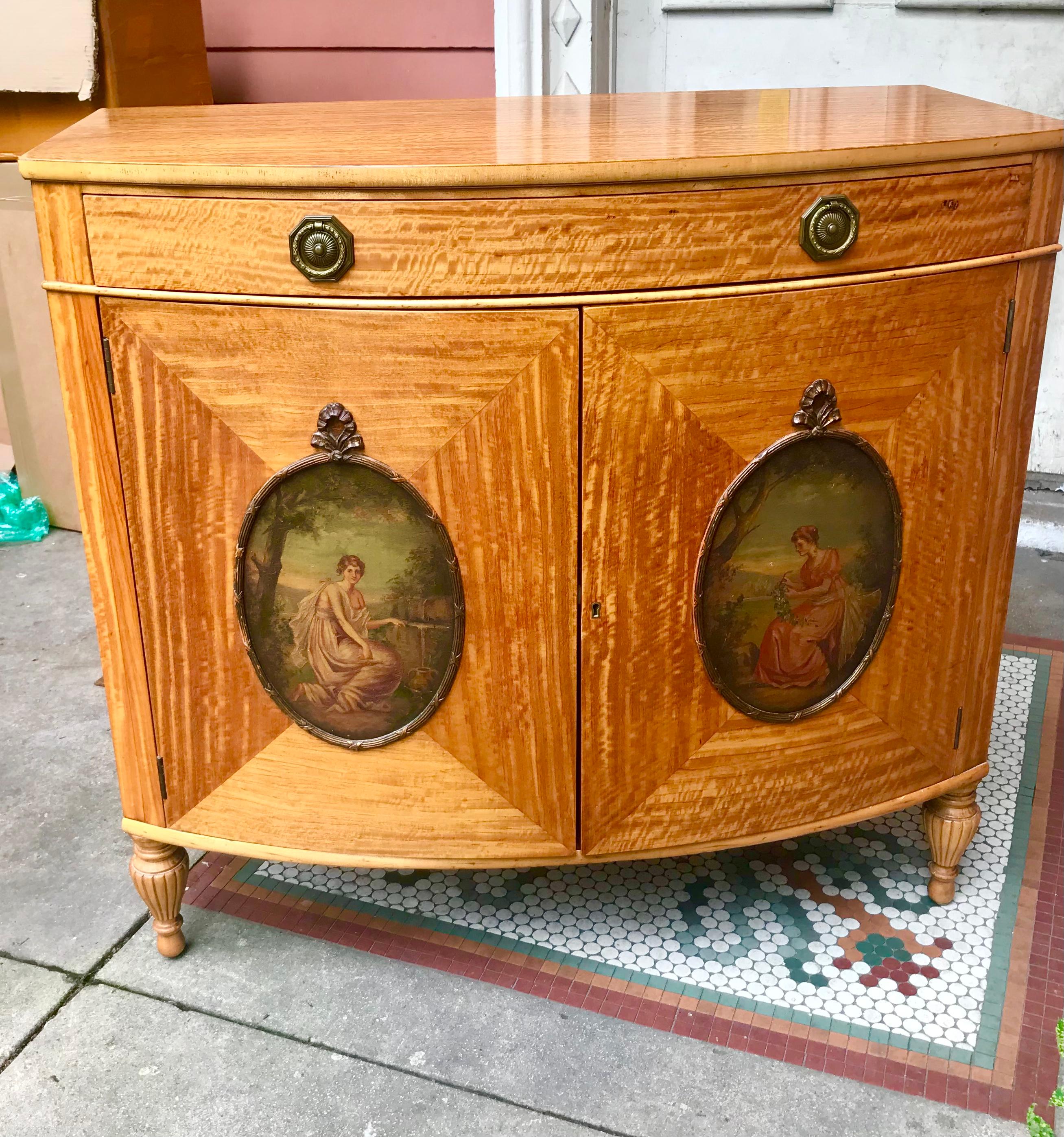 A bowfront cabinet in the Edwardian satinwood style with oval insets of classical ladies in the manner of Adam Painter 
Angelica Kauffman . Presents very well .Beautifully rippled wood graining that changes as you move in almost pristine condition .