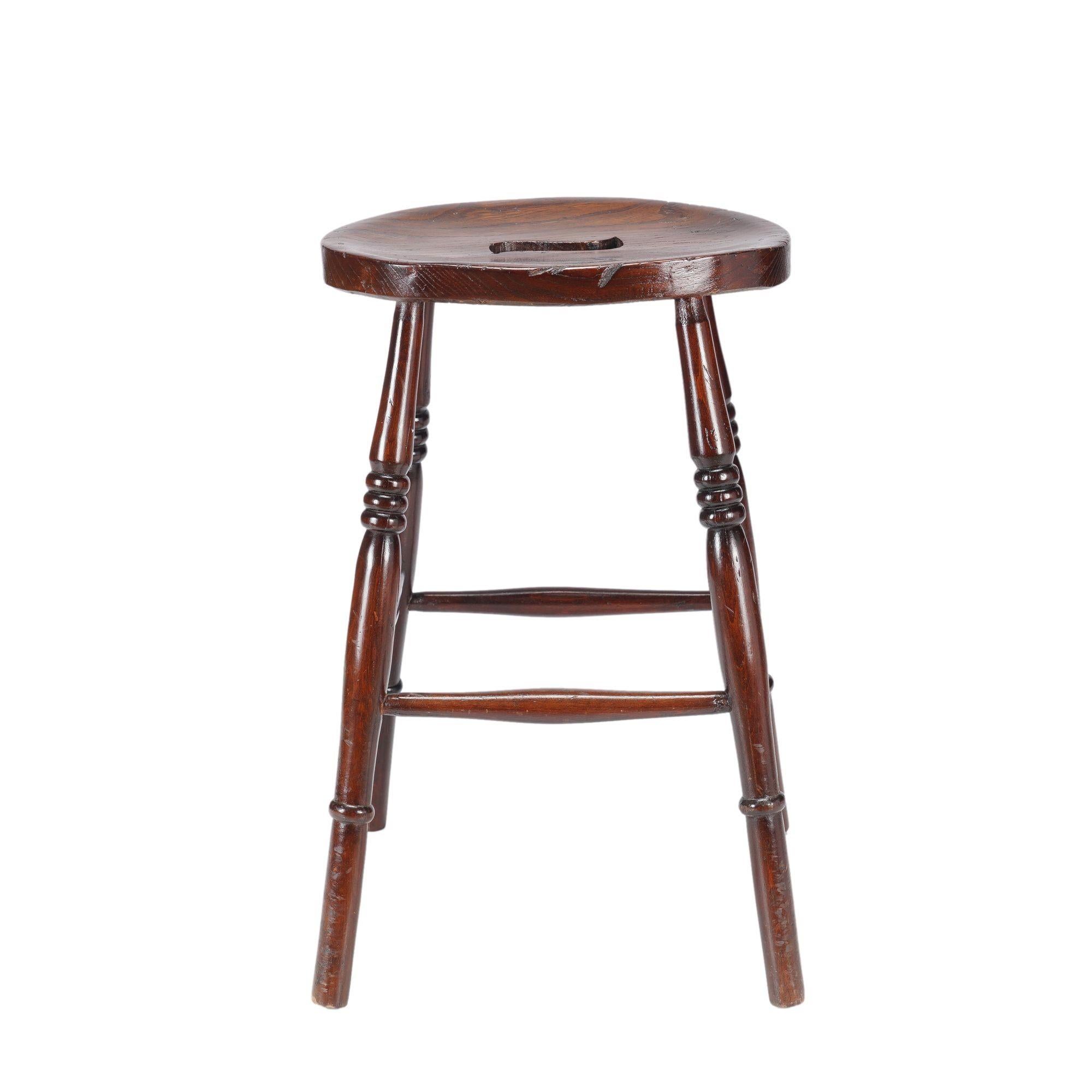English elm pub stool with an oval single board seat centered on a pierced hand grip. The seat is supported by four turned legs which are joined by turned boxed stretchers.

England, circa 1820-40.