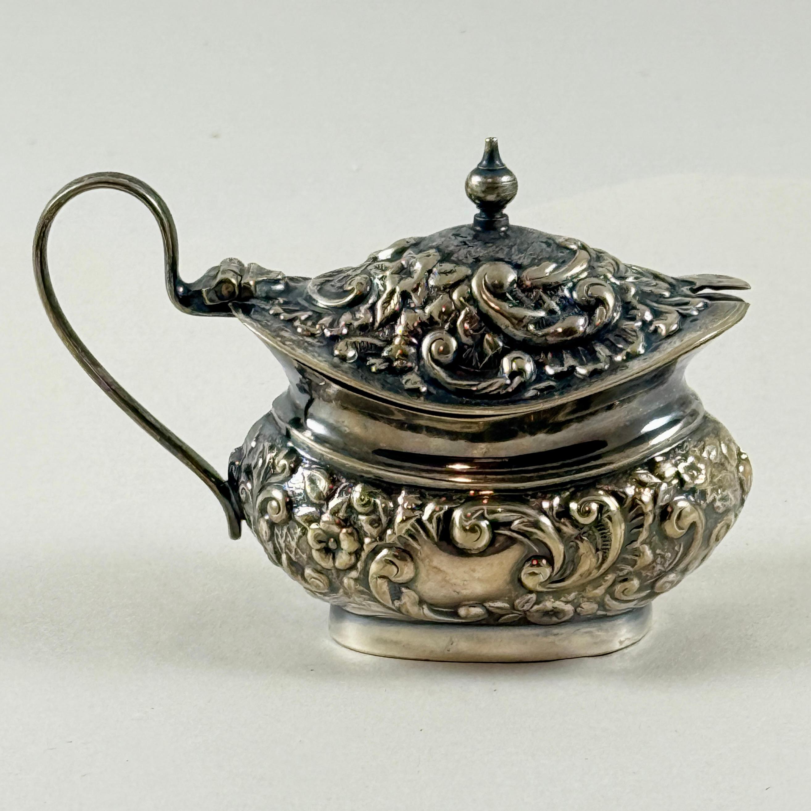 Une salière anglaise en argent EPNS, vers 1890-1910.

Ciselé d'un motif floral et de rinceaux en repoussé, il est muni d'un couvercle à charnière et d'un bouton supérieur.
Doublé d'un porte-sel en plastique bleu. Le couvercle est muni d'une fente