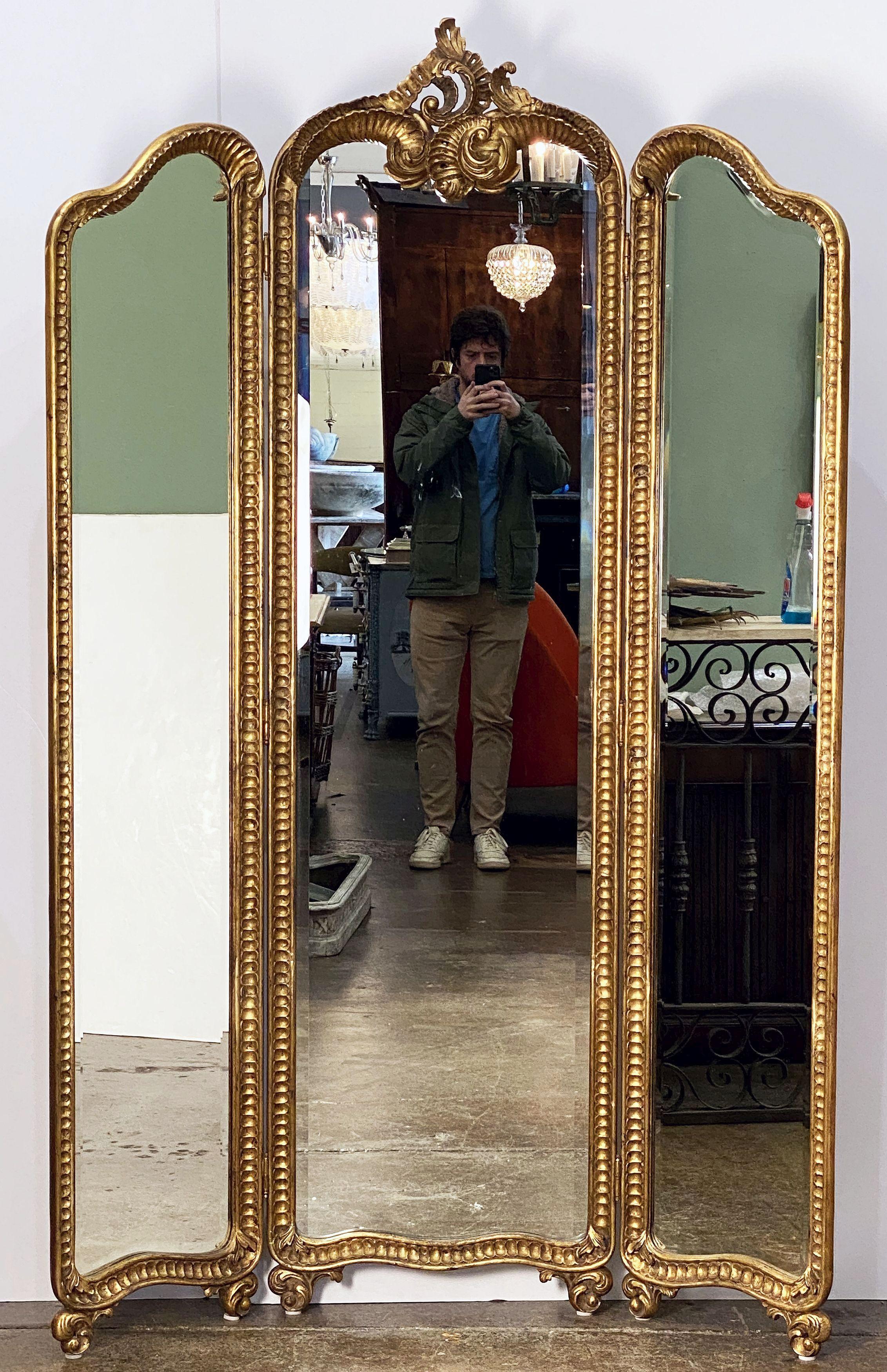 A fine English three-panel folding floor-standing dressing mirror, circa 1920, featuring a carved giltwood center frame with two removable side panels - each embellished with a Rococo design and shaped beveled mirror glass, standing on stylized