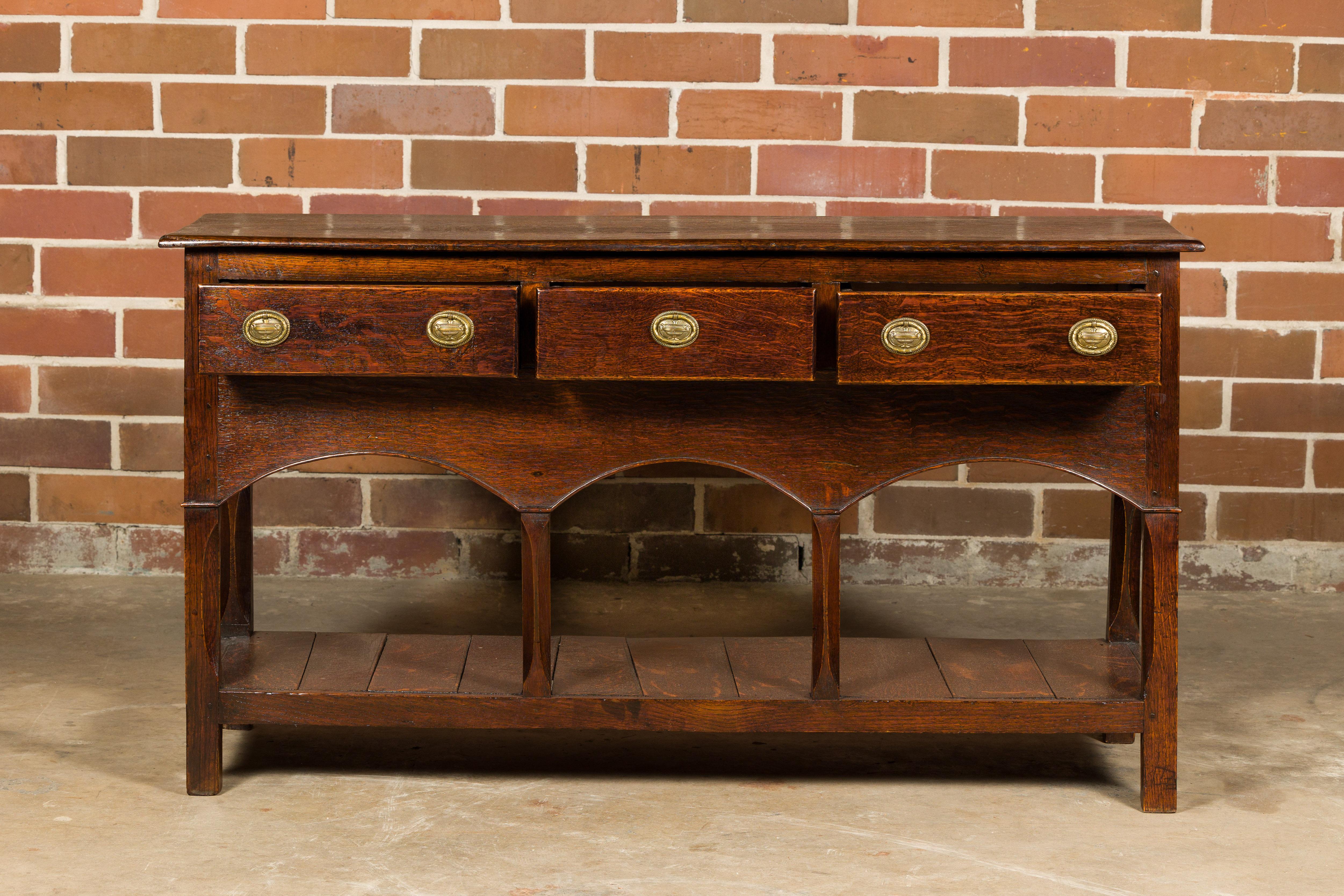 An English George III period oak dresser base from circa 1800 with three drawers, arching accents on the apron a low shelf. This English George III period oak dresser base, crafted around 1800, is a timeless testament to classic design and sturdy