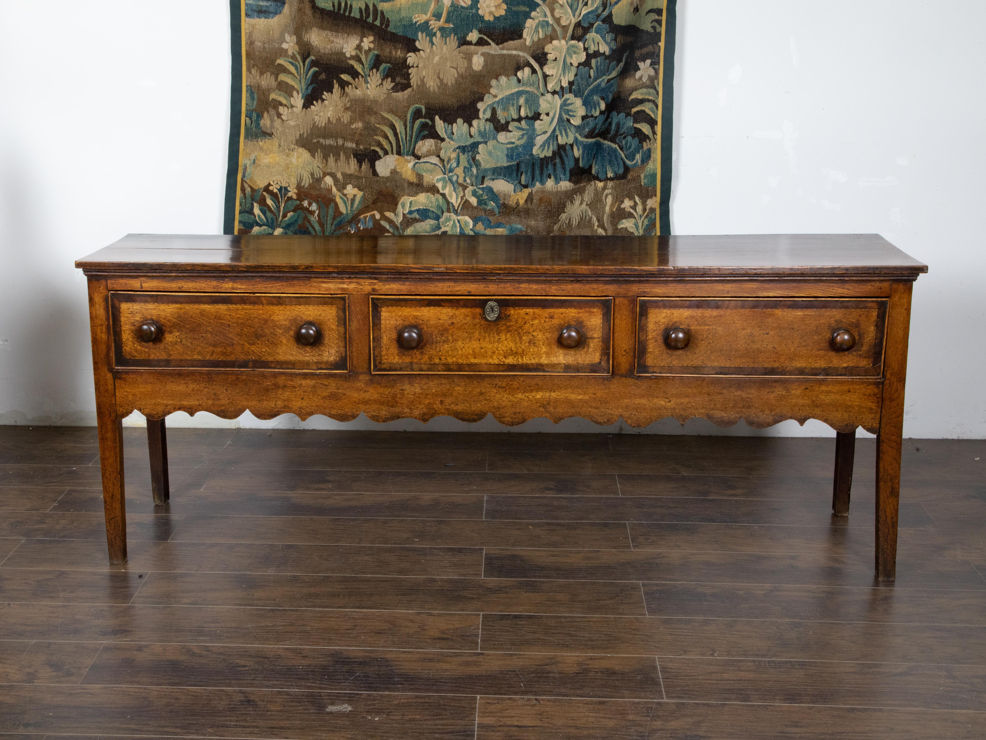 An English George III period oak dresser base from the early 19th century, with three drawers, inlay, scalloped apron and straight legs. Created in England during the reign of King George III in the early years of the 19th century, this oak dresser