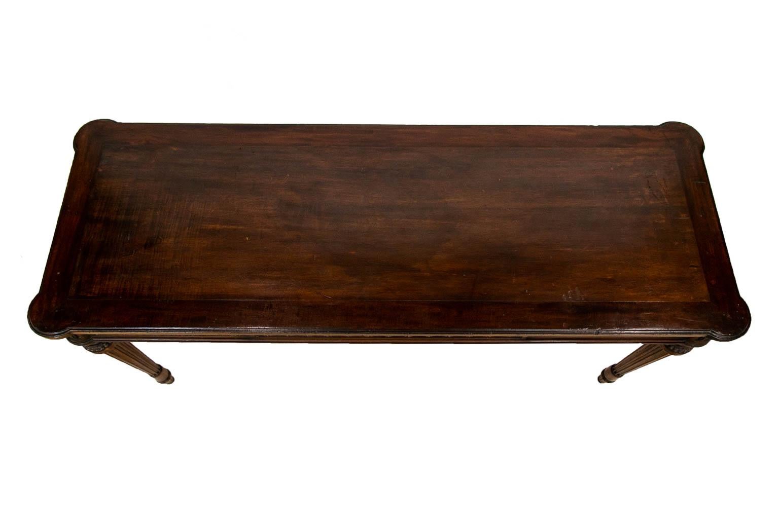 The top of this mahogany center table has a three inch border framing the center panel and has shaped edging. The front and back sides have a repeating interlaced vine and rosette panel. The ends have a recessed raised panel. The four legs each have