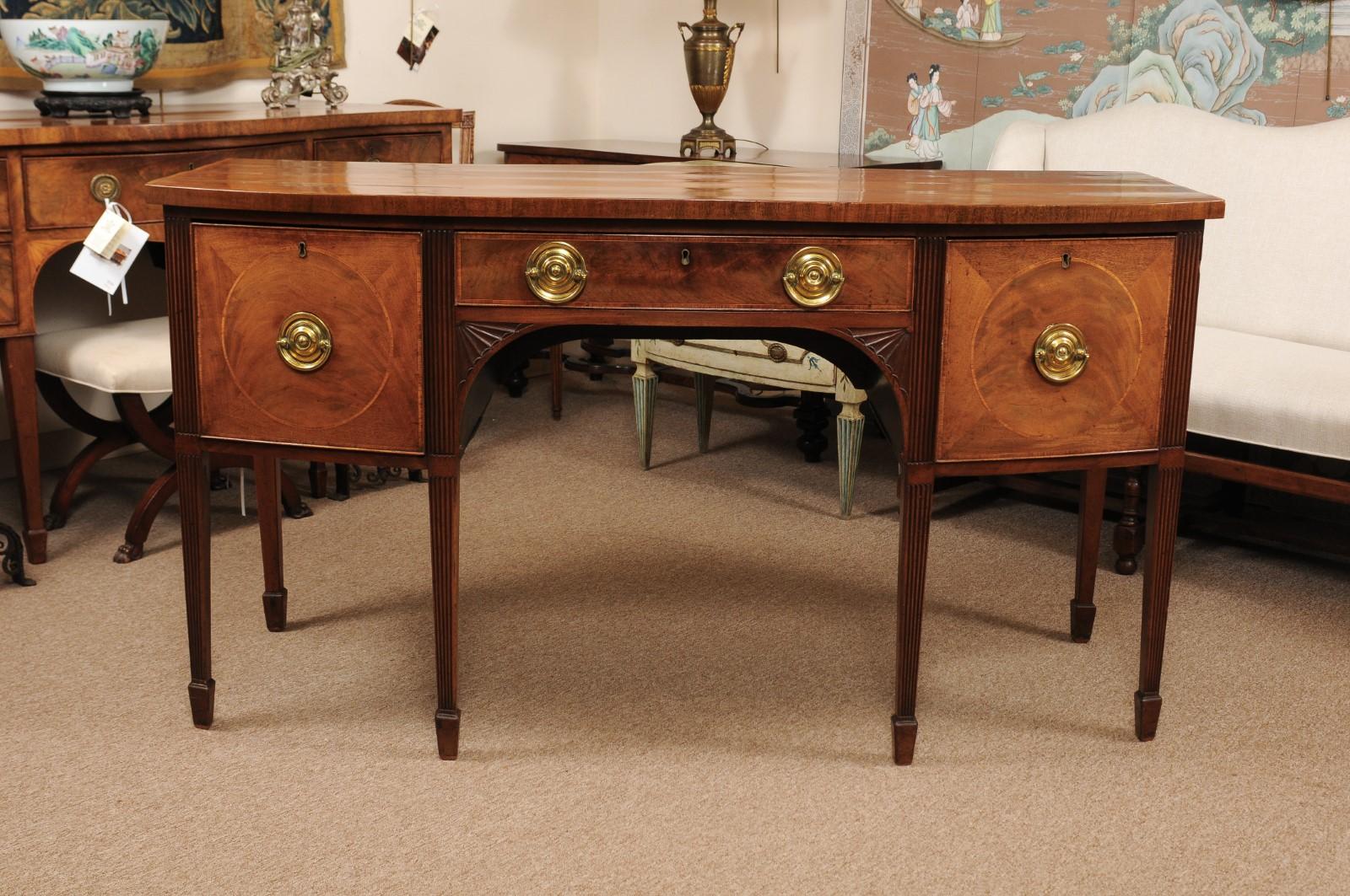 An early 19th century George III English mahogany bow-front sideboard featuring rosewood crossbanding and string boxwood inlay on drawers, fan carved brackets below ending on reeded legs and spade feet.

     