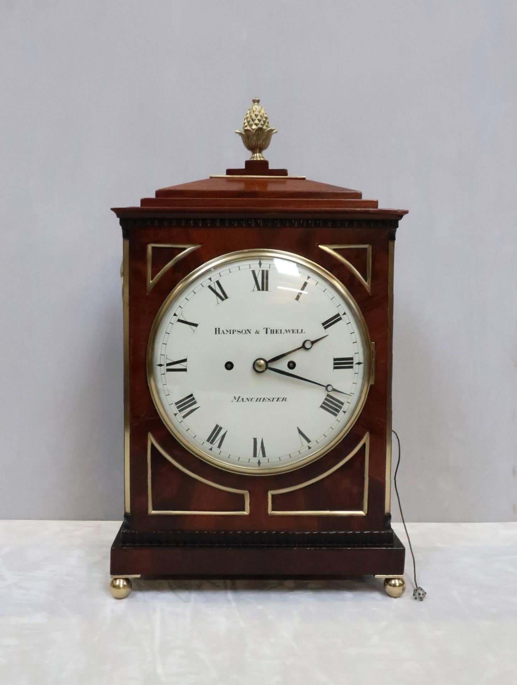 A very good quality English George III figured mahogany chamfer top bracket or table clock with ebony mouldings, inset corner panels and brass moulded edges to the front of the case stood on brass ball feet finished with a brass pineapple finial to