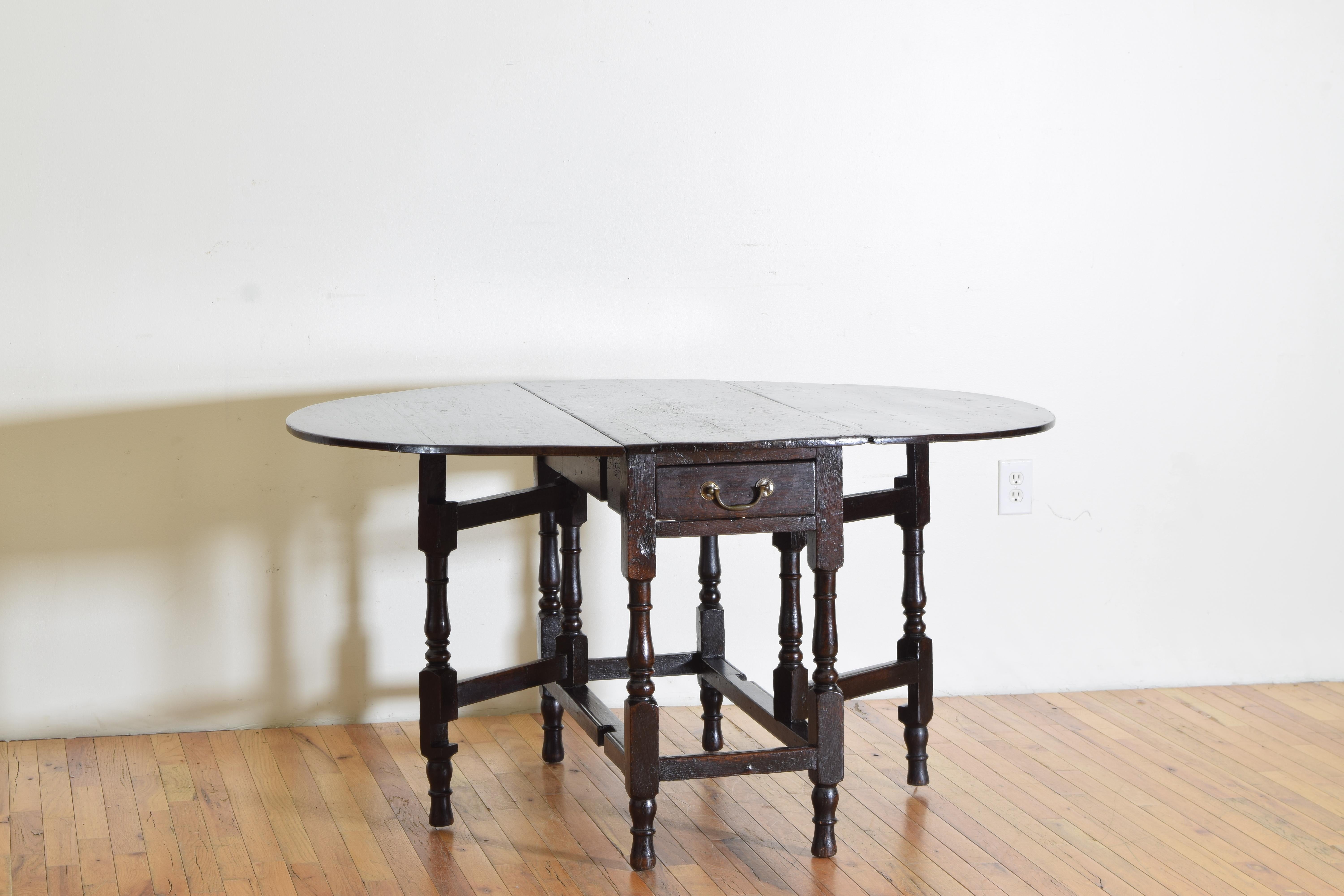 Having, when fully opened, a top between oval and round resting on a metamorphic base featuring bobbin turned legs with block stretchers, the side legs hinged to allow support for the folding top, containing two deep drawers with original brass