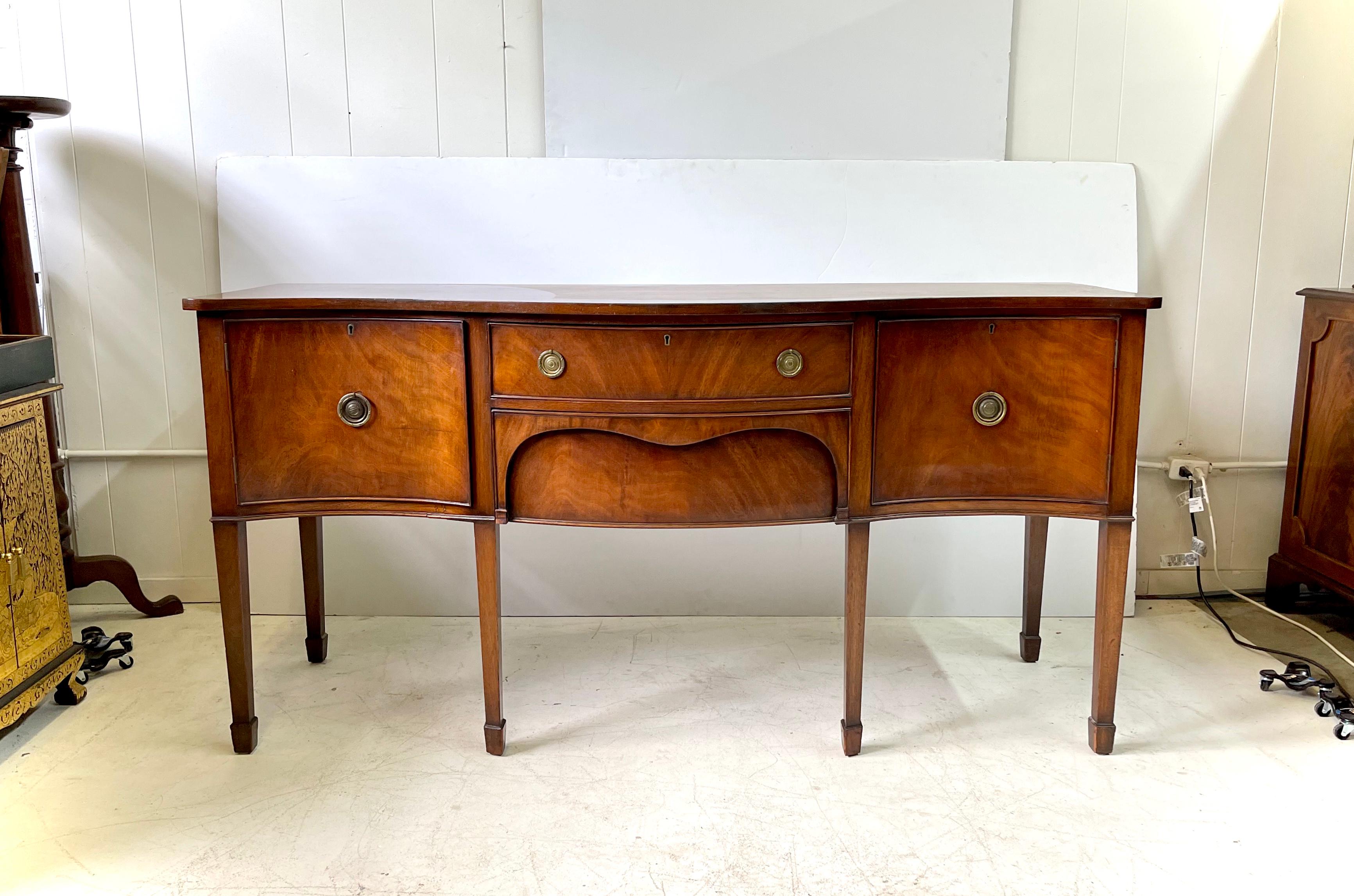 19th century English sideboard, serpentine in shape and constructed of flame mahogany, made in the manner of George III. A shaped one board top rests on a case with beaded cabinets flanking a fitted felt-lined silver drawer over a recessed hidden