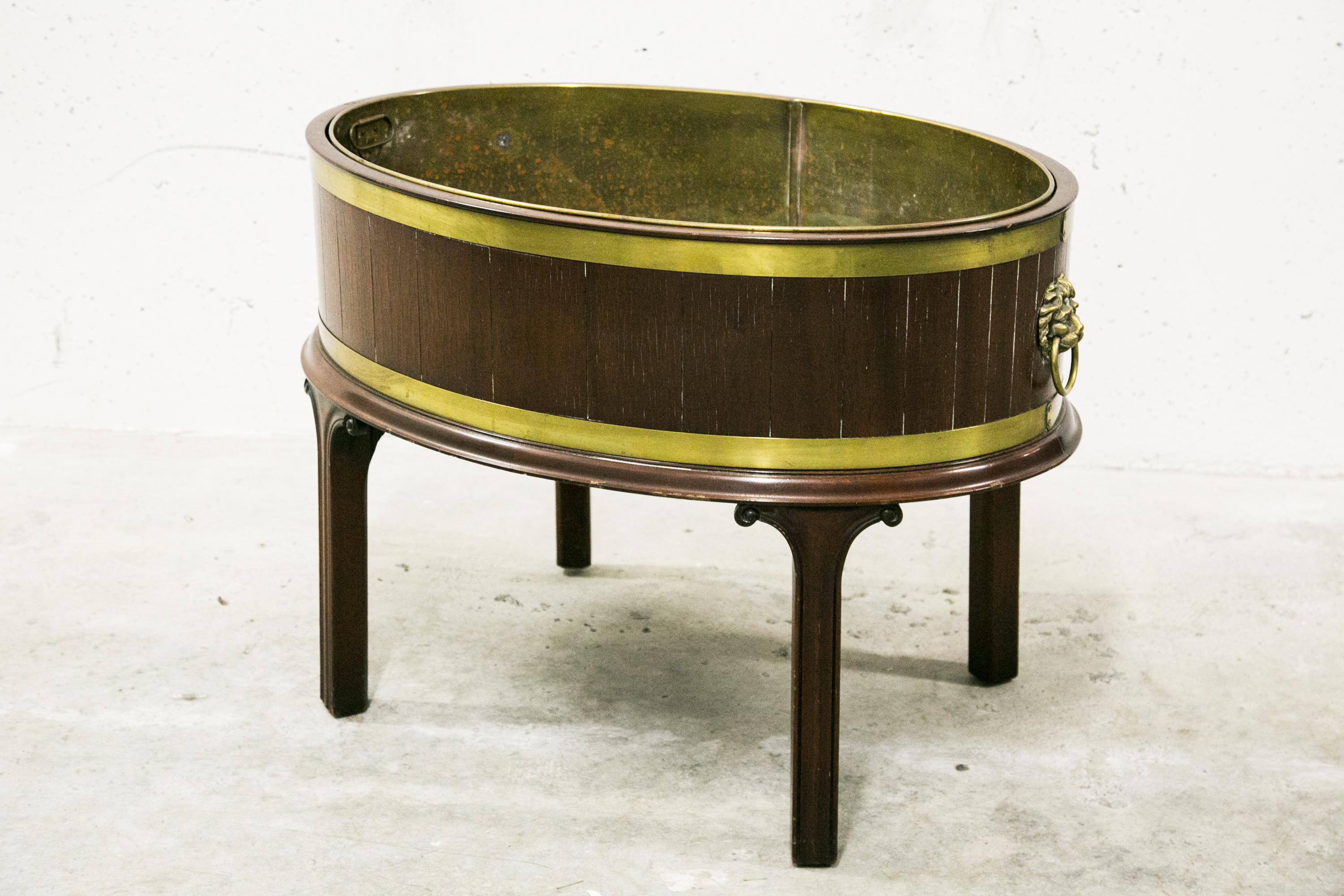 This is a classic oval English George III style period appropriate mahogany wine cooler. The exterior walls are constructed in vertical plank-forms bound together with upper and lower brass straps. The interior is metal lined. Brass lion’s head