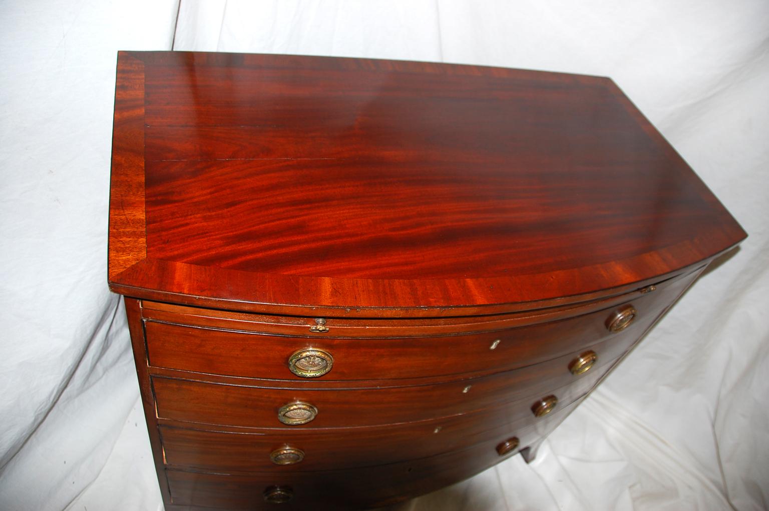 English Georgian Hepplewhite period mahogany bowfront chest of four graduated drawers with dressing slide. The graceful shaped skirt has a sunburst inlay in satinwood and boxwood which sets this chest apart from the normal. This small chest of