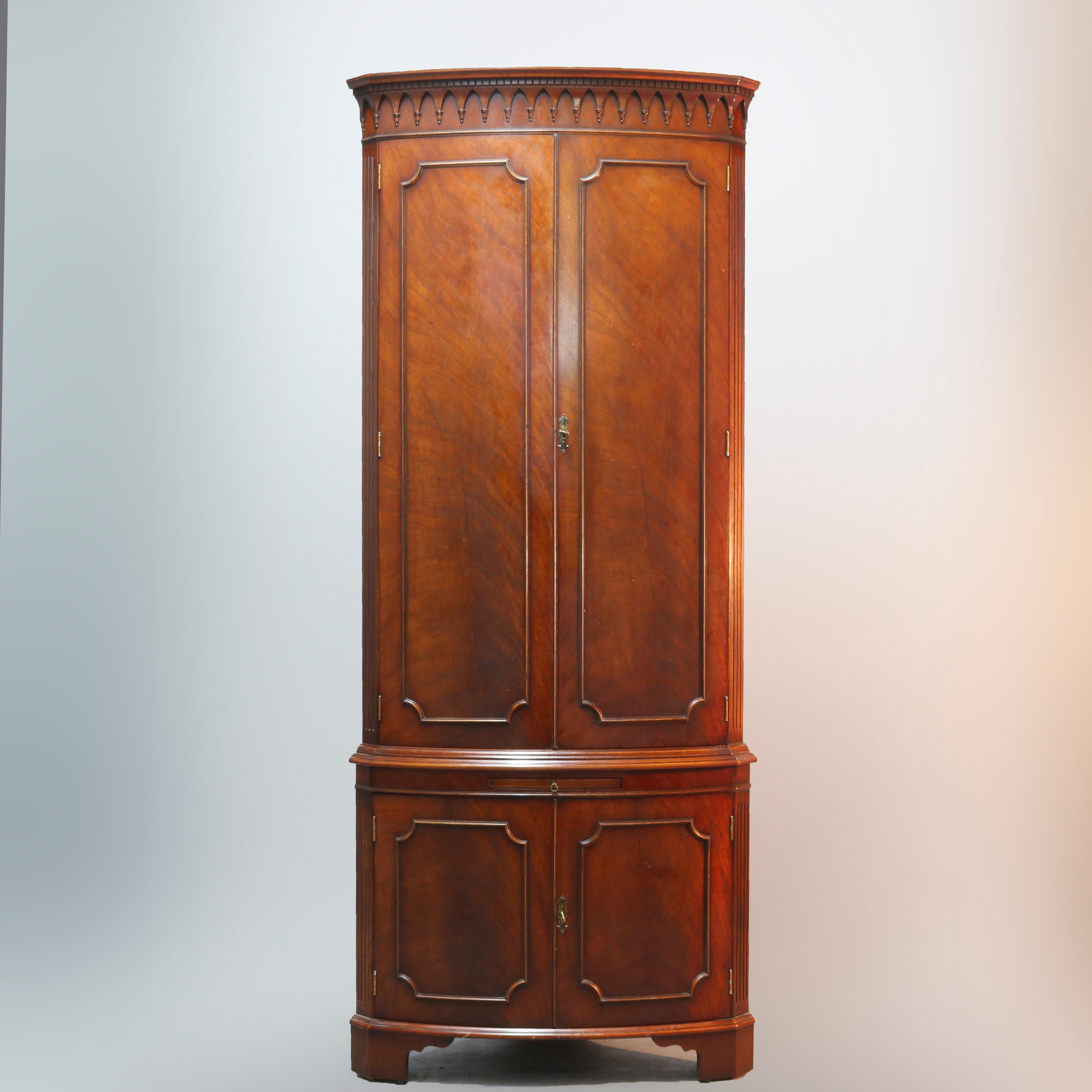 An English George III style corner cabinet offers flame mahogany construction with double door upper having arched panels and opening to lighted shelved and mirrored interior, surmounting double door lower cabinet and raised on bracket fee, 20th
