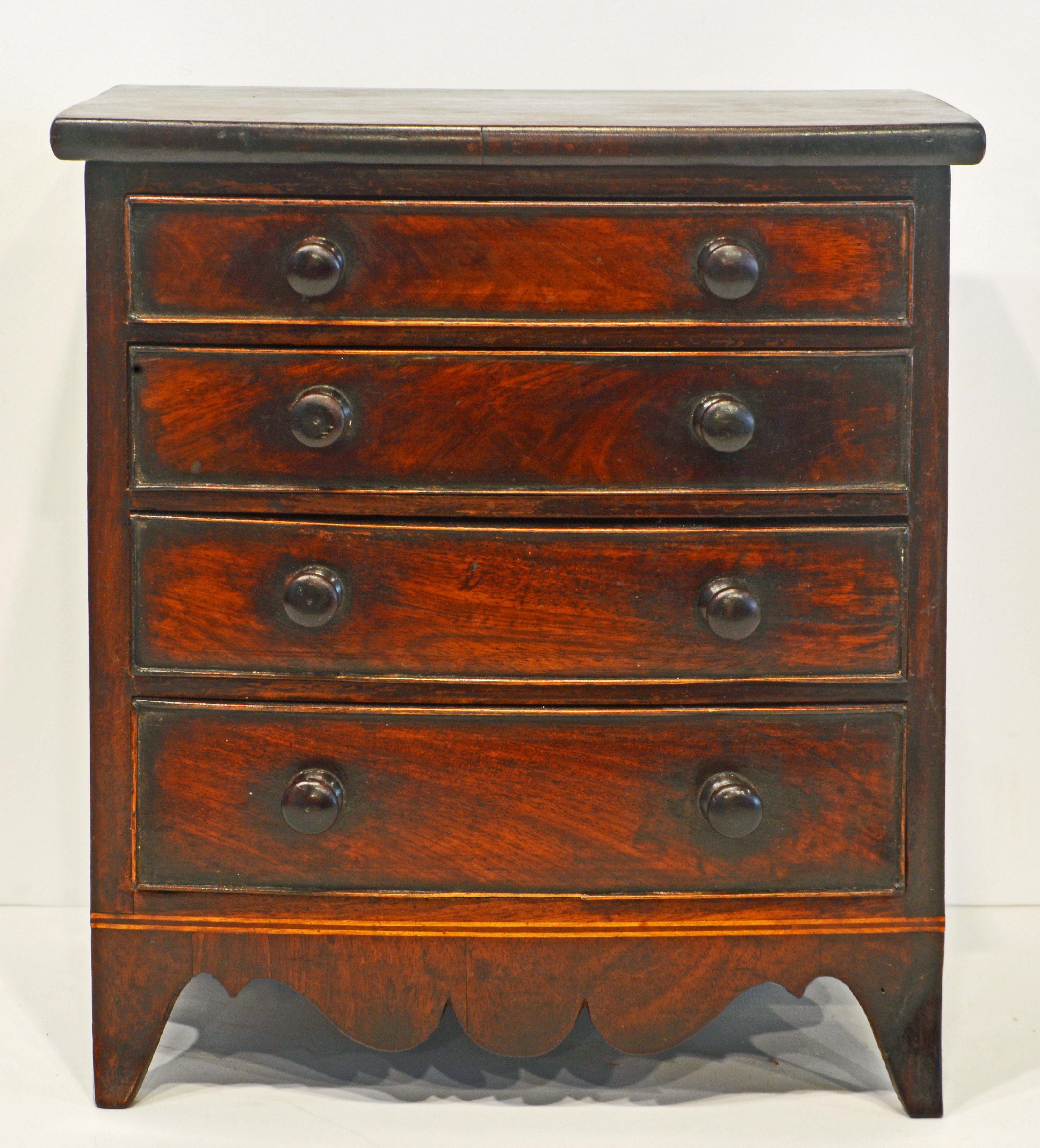 This very distinguished Georgian miniature mahogany bow front chest of drawers dating to the early 19th century features a mahogany top above four graduated long drawers retaining their original wooden knobs and a boldly scalloped apron flanked by