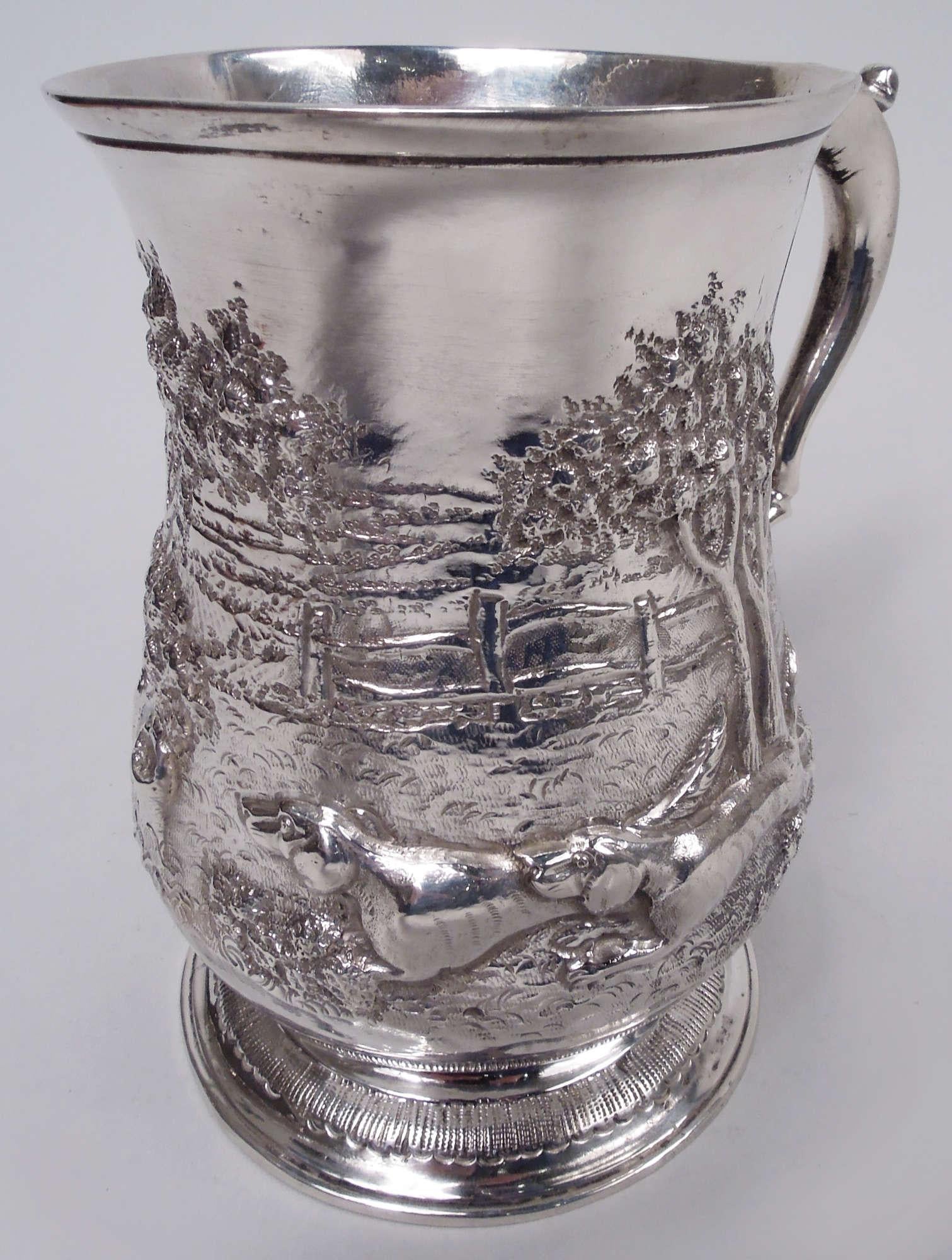 Mug en argent de George II. Fabriqué par Samuel Welles à Londres en 1759. Coupe balustre avec poignée à double volute coiffée d'une feuille. Pied surélevé avec bordure de feuilles estampées. Scène bucolique ciselée et gravée avec des chiens courants