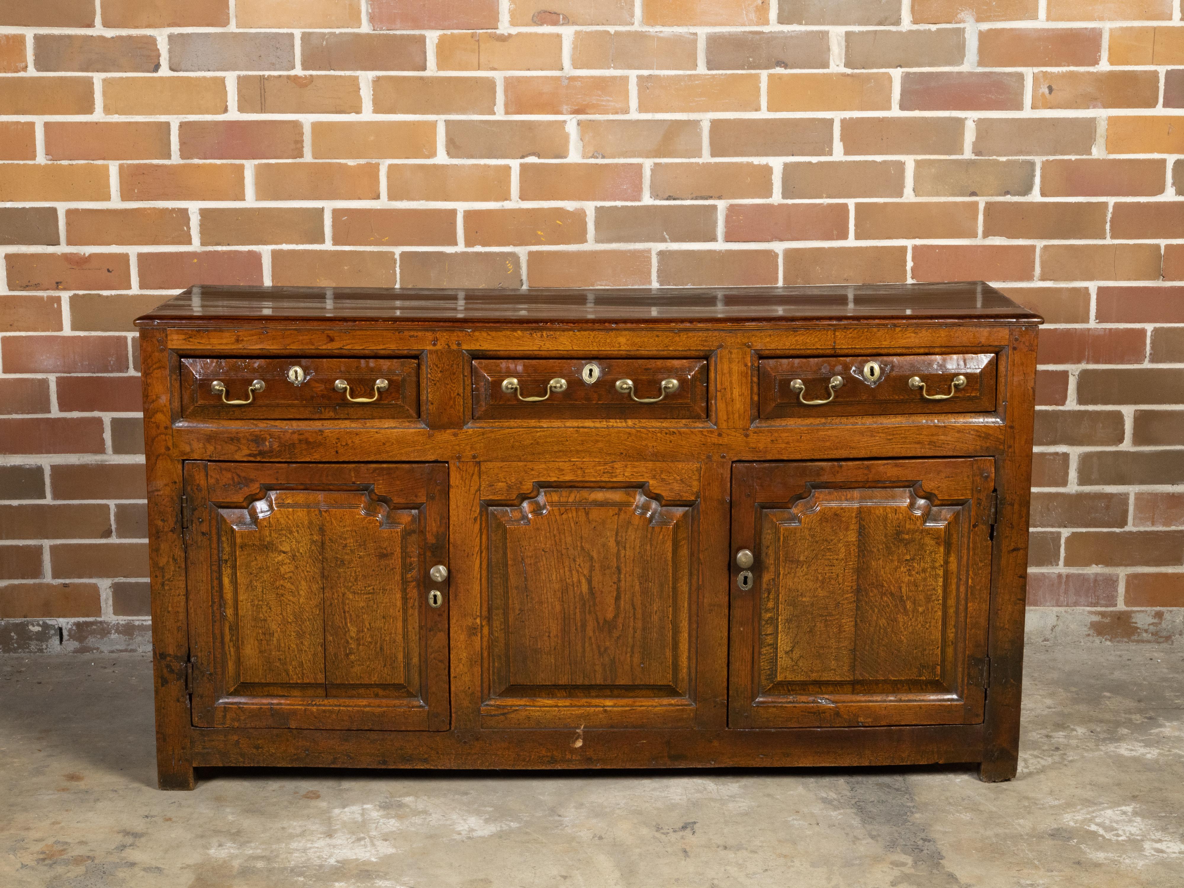 An English Georgian period oak dresser base from the early 19th century with three drawers, two doors, carved panels and brass hardware. Created in England during the Georgian period in the early years of the 19th century, this oak dresser base